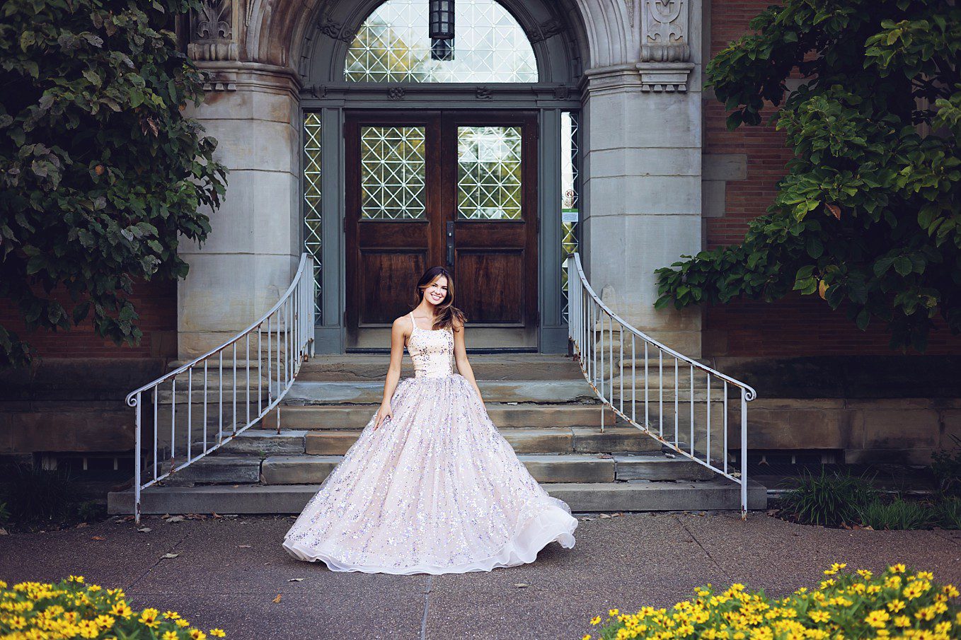 image of a senior girl wearing a cream colored gown. She's standing in front of an old stone building at Phipps Conservatory in Pittsburgh. There are yellow daisies at her feet