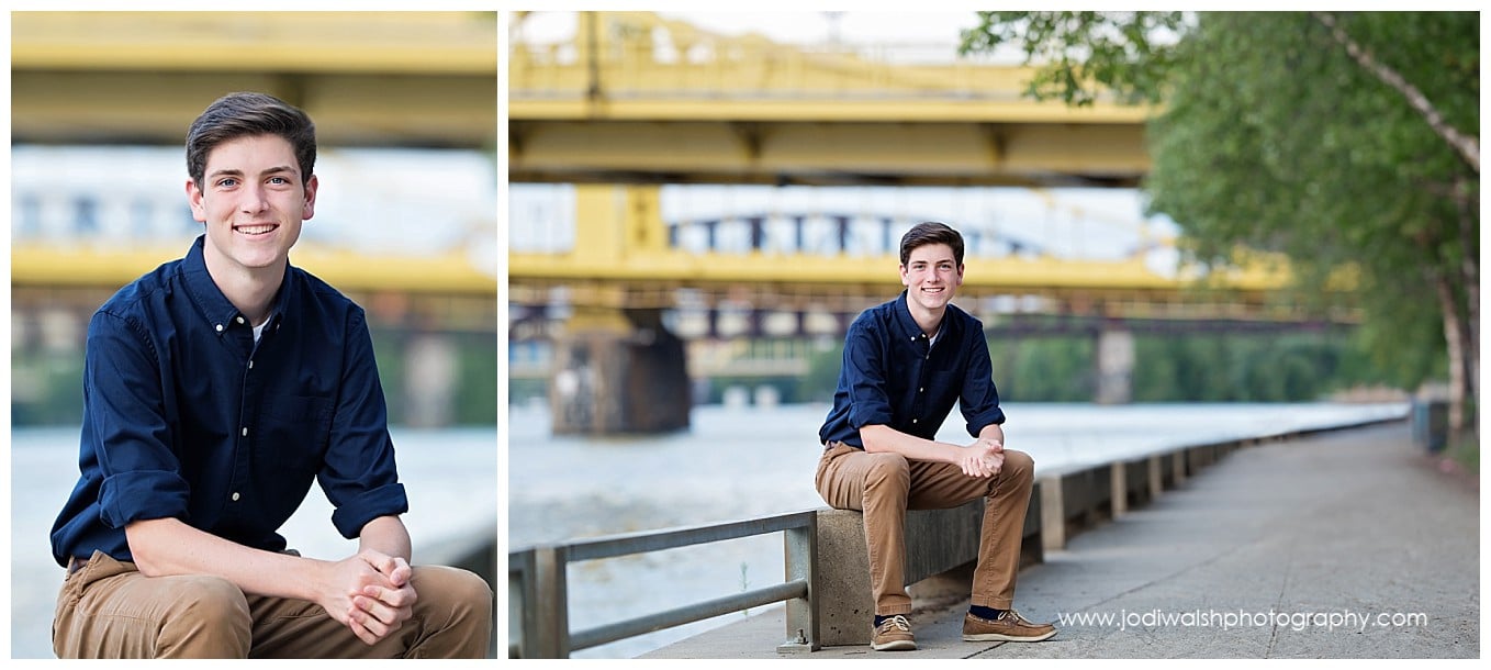 images of a senior guy wearing a navy blue button down shirt, sitting on a railing by the river with a yellow bridge behind