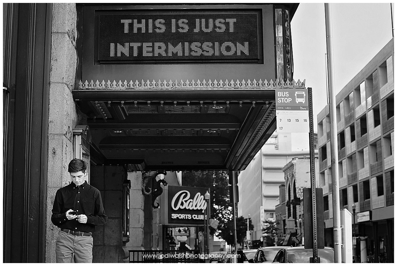 black and white image of a Pittsburgh senior young man looking at his cell phone while standing under the Byham theater marquee that reads This is Just Intermission