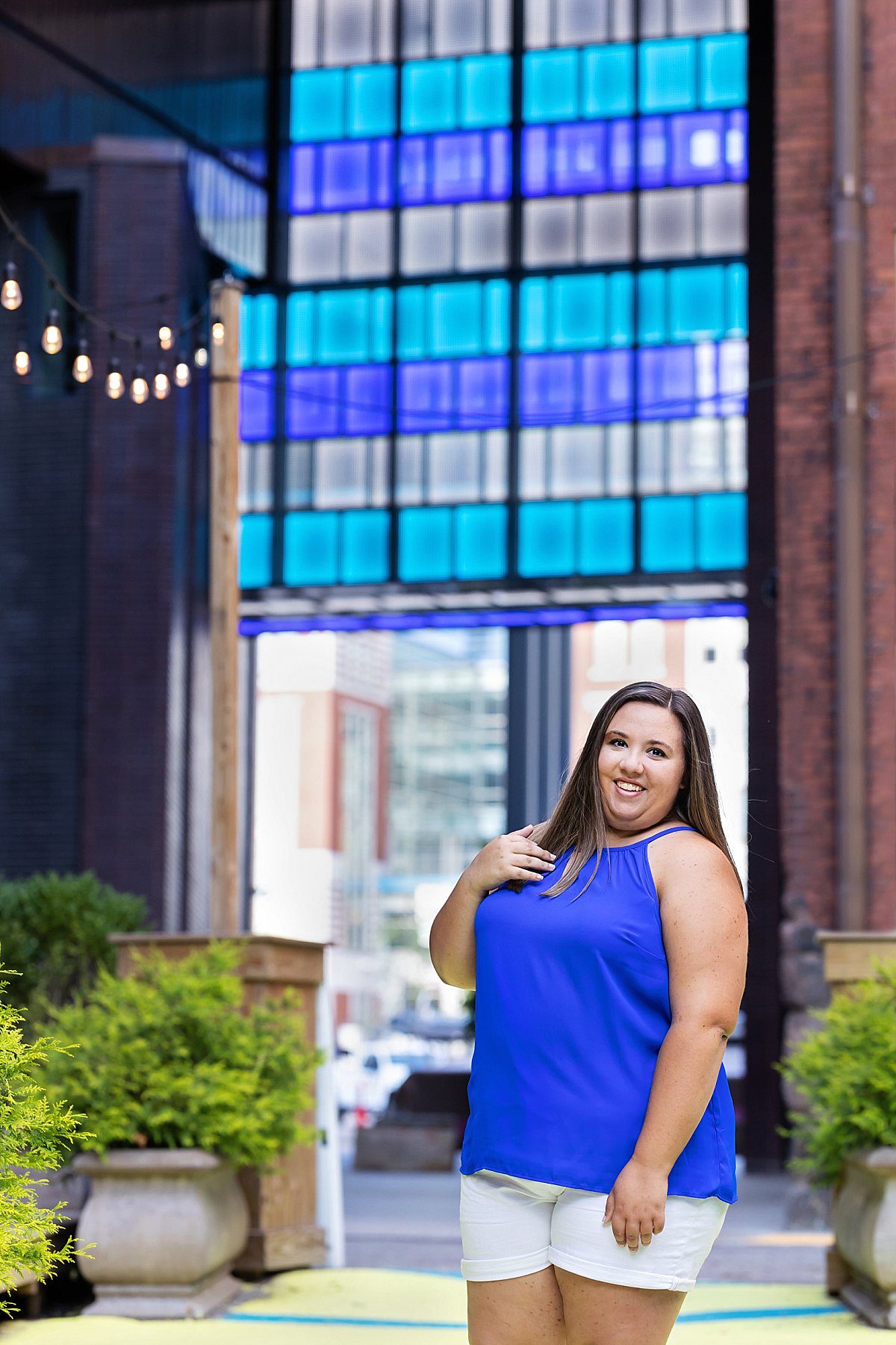 image of a senior girl in a blue top with a bright blue light installation above her. She's smiling as she stands in an outdoor garden in downtown Pittsburgh.