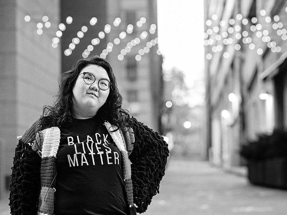 black and white images of a girl wearing a Black Lives Matter t-shirt. She standing in front of an alley with lights hanging behind her and she's looking up