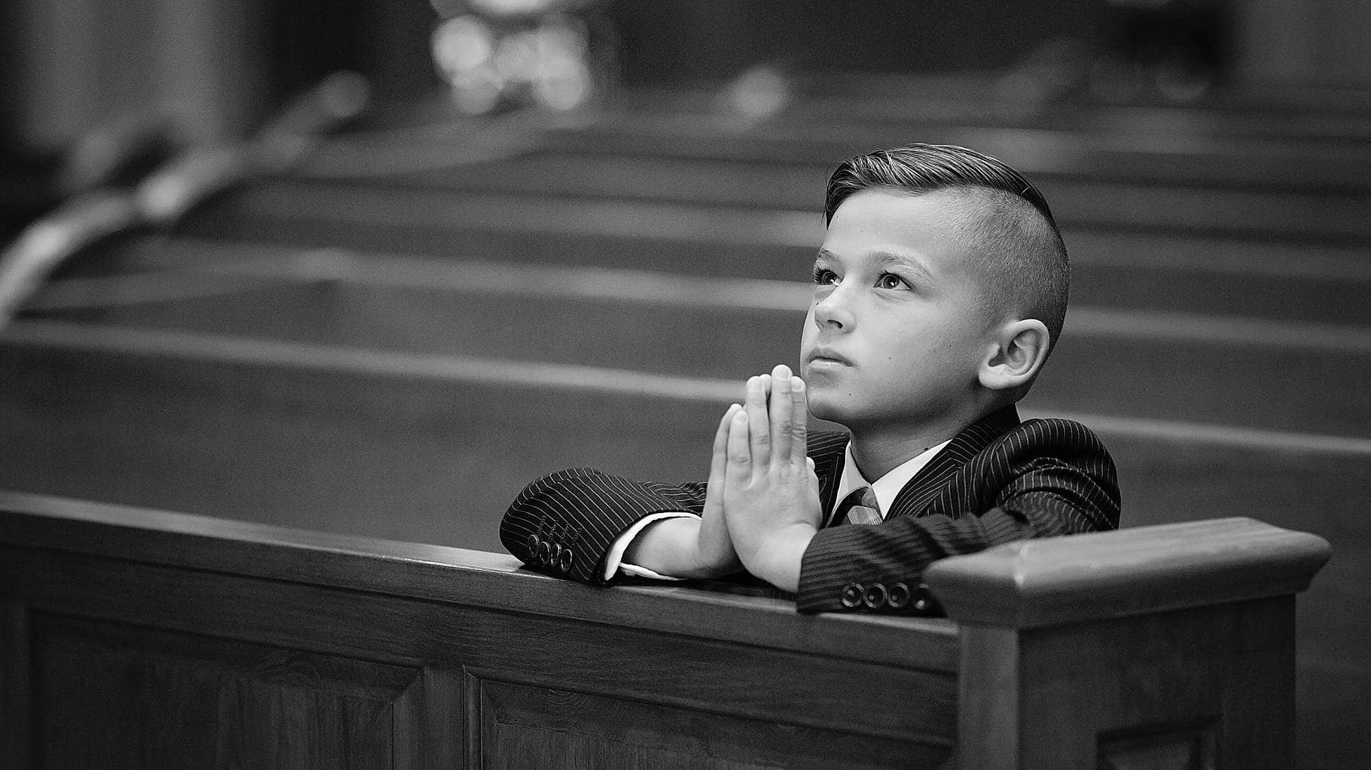 black and white image of a boy in a church pew with his hands folding in prayer