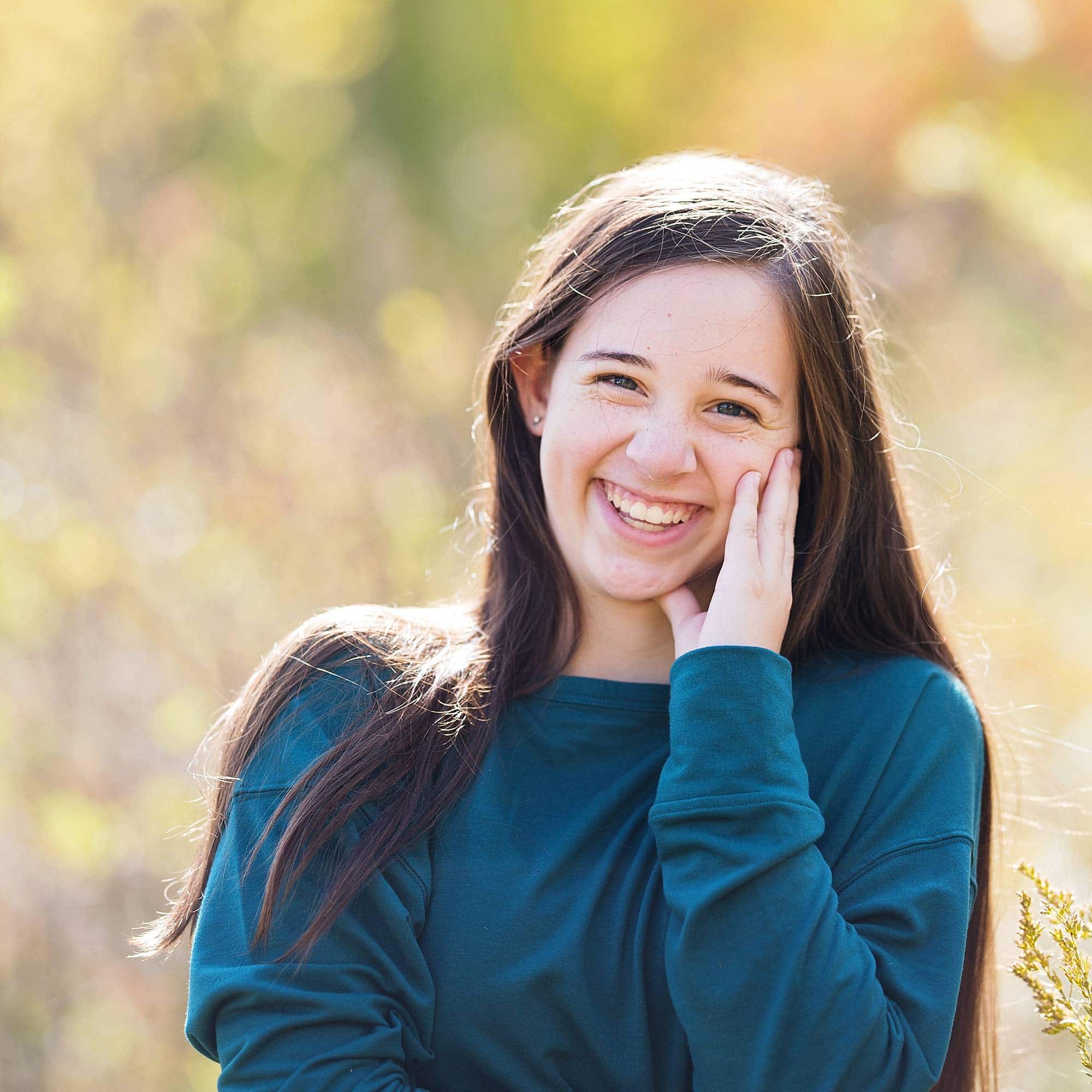 image of a teen girl with long dark hair, wearing a blue green sweater, standing in a golden field grinning with her hand up by her cheek