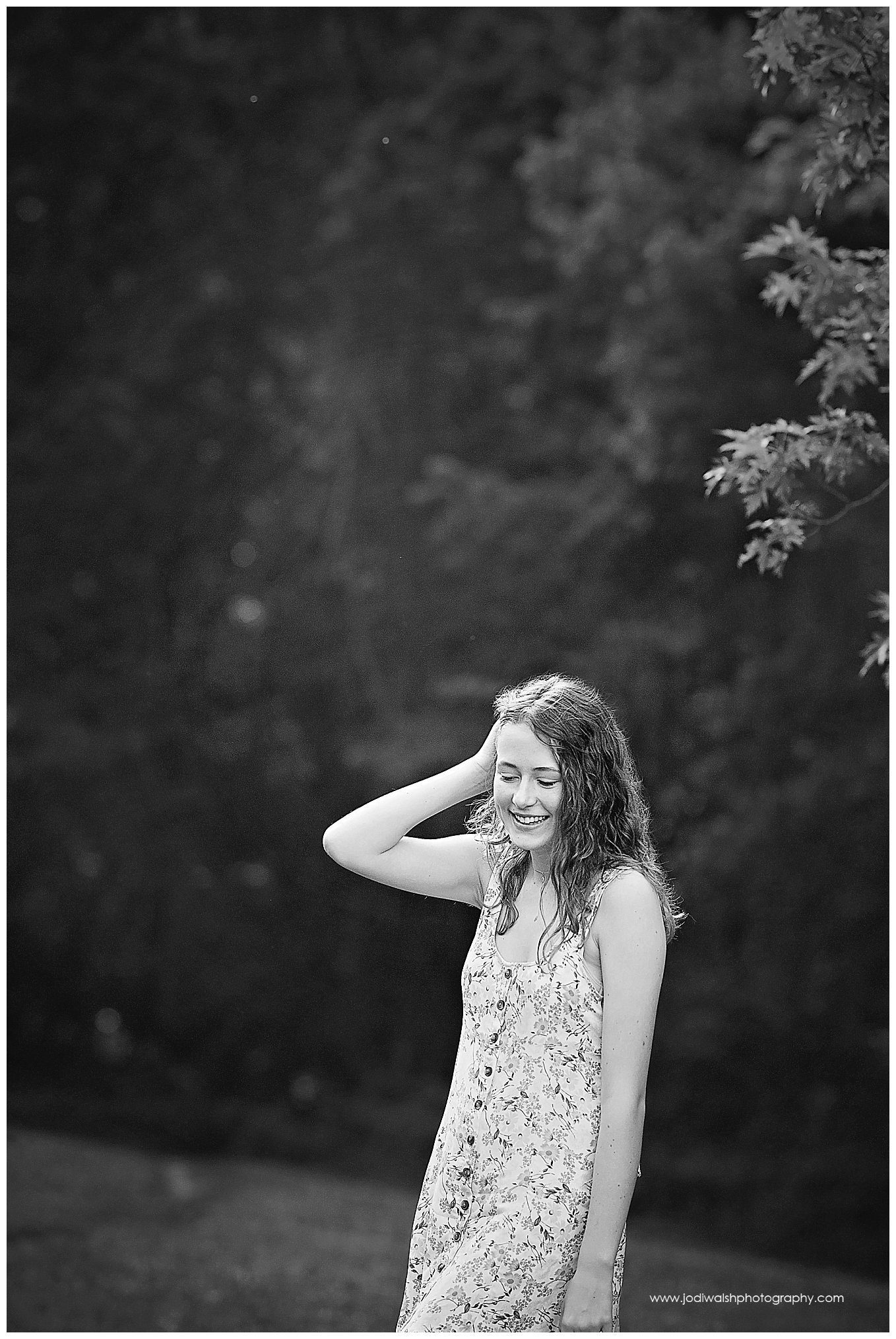 black and white images of a senior girl in a wooded clearing in North Park.  She's wearing a white dress and is looking down as she pushes her hair behind her ear.