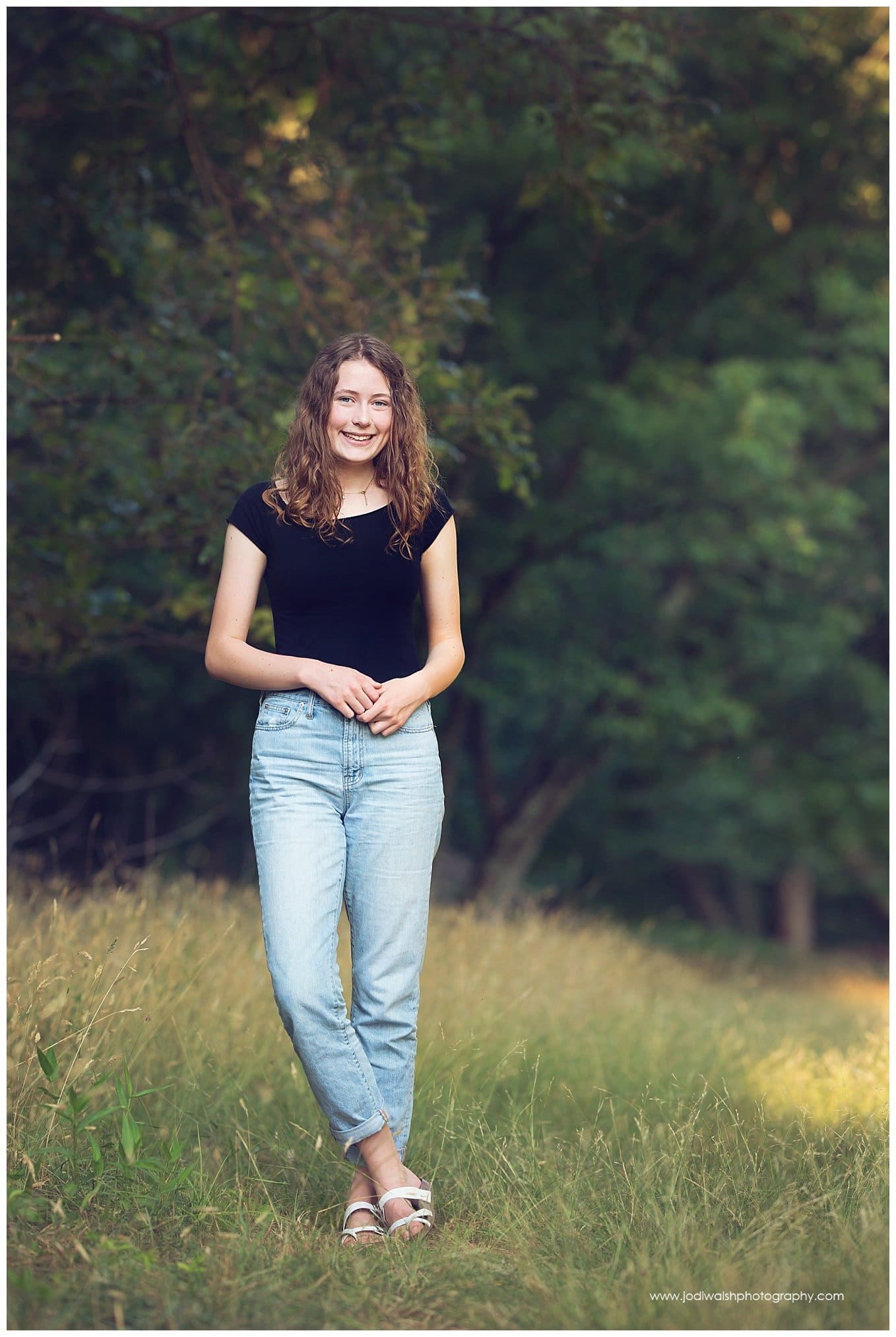 image of a senior girl in North Park.  She's wearing bluejeans and a black t-shirt and she's standing in a field of tall grasses at the edge of a wood.