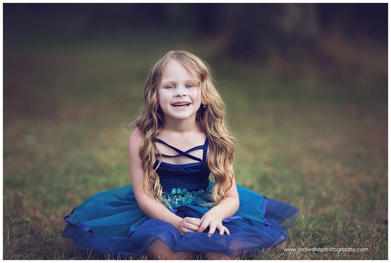 image of a little girl wearing a blue tutu. She's sitting in the grass smiling and laughing.