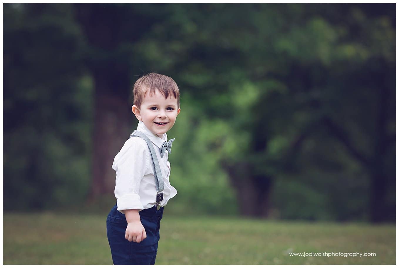 image of a little boy wearing a bow tie and suspenders.  He's smiling at the camera while standing in a field in North Park.