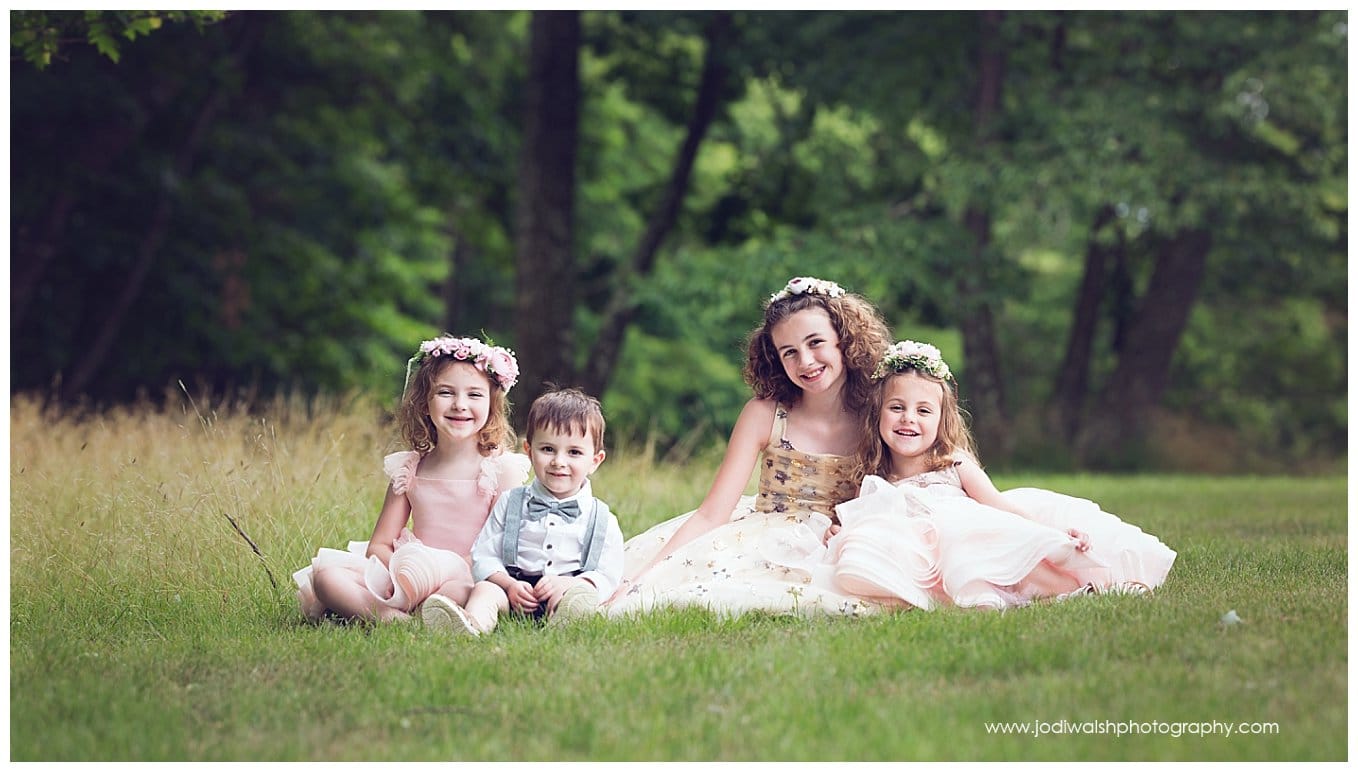 image of three sisters and a little brother, sitting in the grass together. The sisters are wearing princess dresses and flower crowns. The little brother is wearing a bow tie and suspenders/