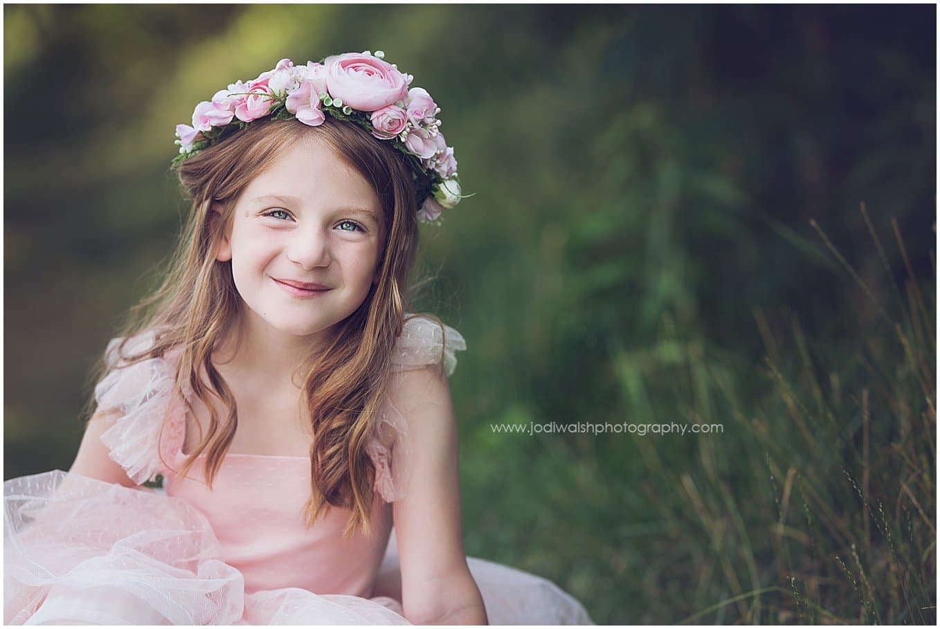 image of a little girl wearing a pink flower crown and a pink princess dress. She's sitting outside in the grass and smiling.
