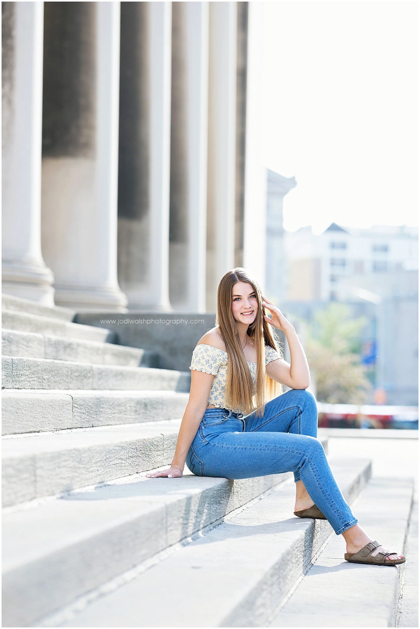 image of a senior girl wearing jeans and a flower top, sitting on the steps of the Mellon Institute in Oakland.