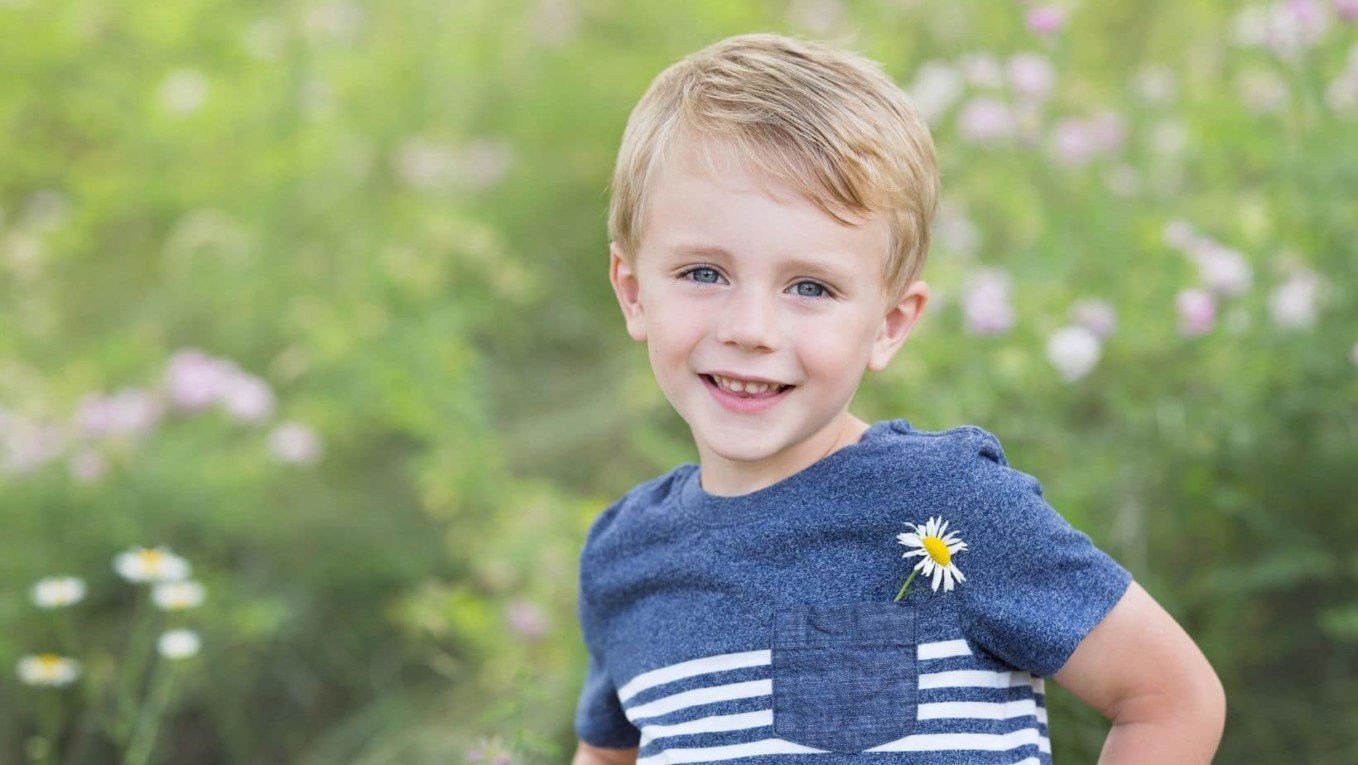little boy with blond hair and blue eyes standing on a hillside with wildflowers. He has a daisy in his shirt pocket.
