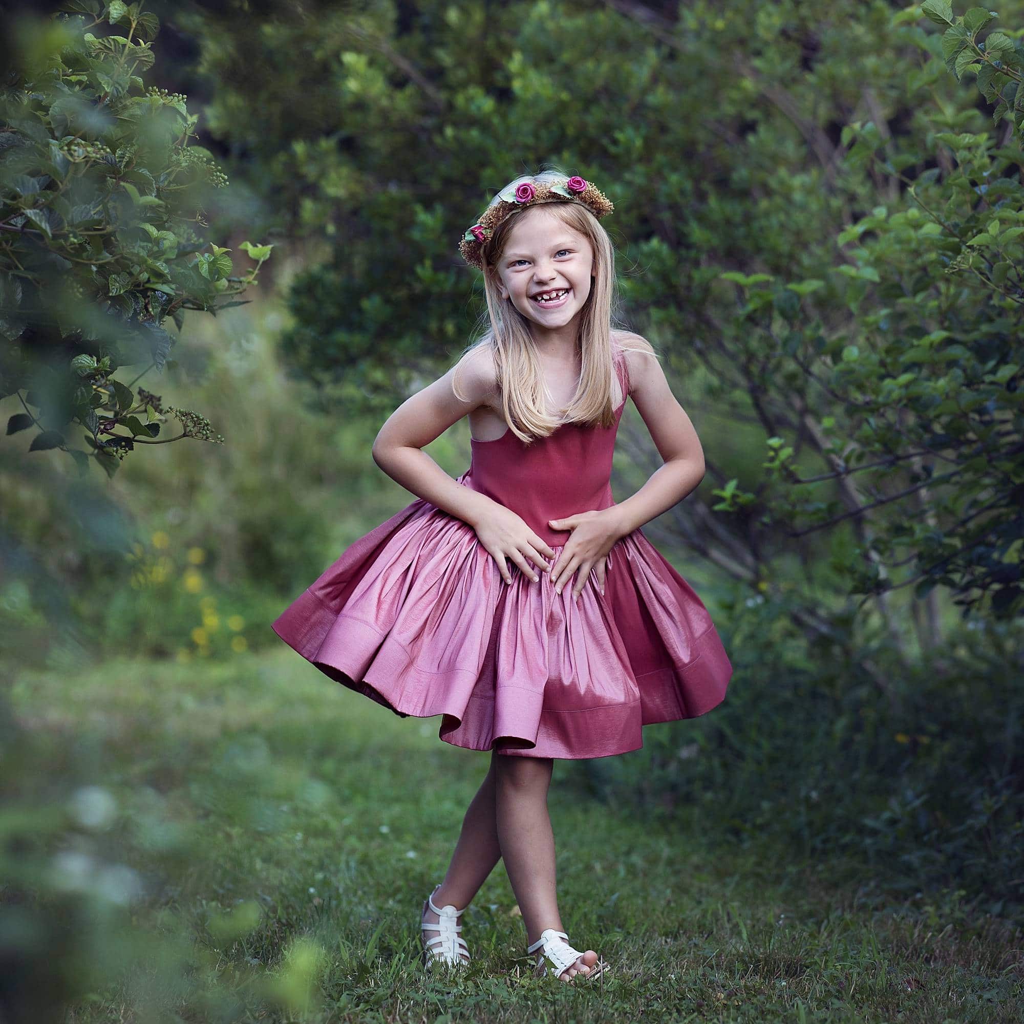 little girl wearing a pink short tutu dress and flower crown. she's smiling while standing near some tall bushes.
