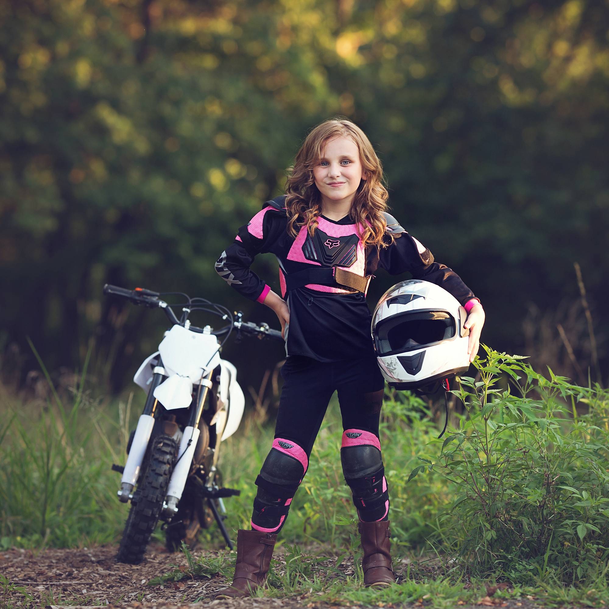 little girl with a white dirt bike. She's wearing a pink and black racing outfit and is holding a white helmet on her hip.
