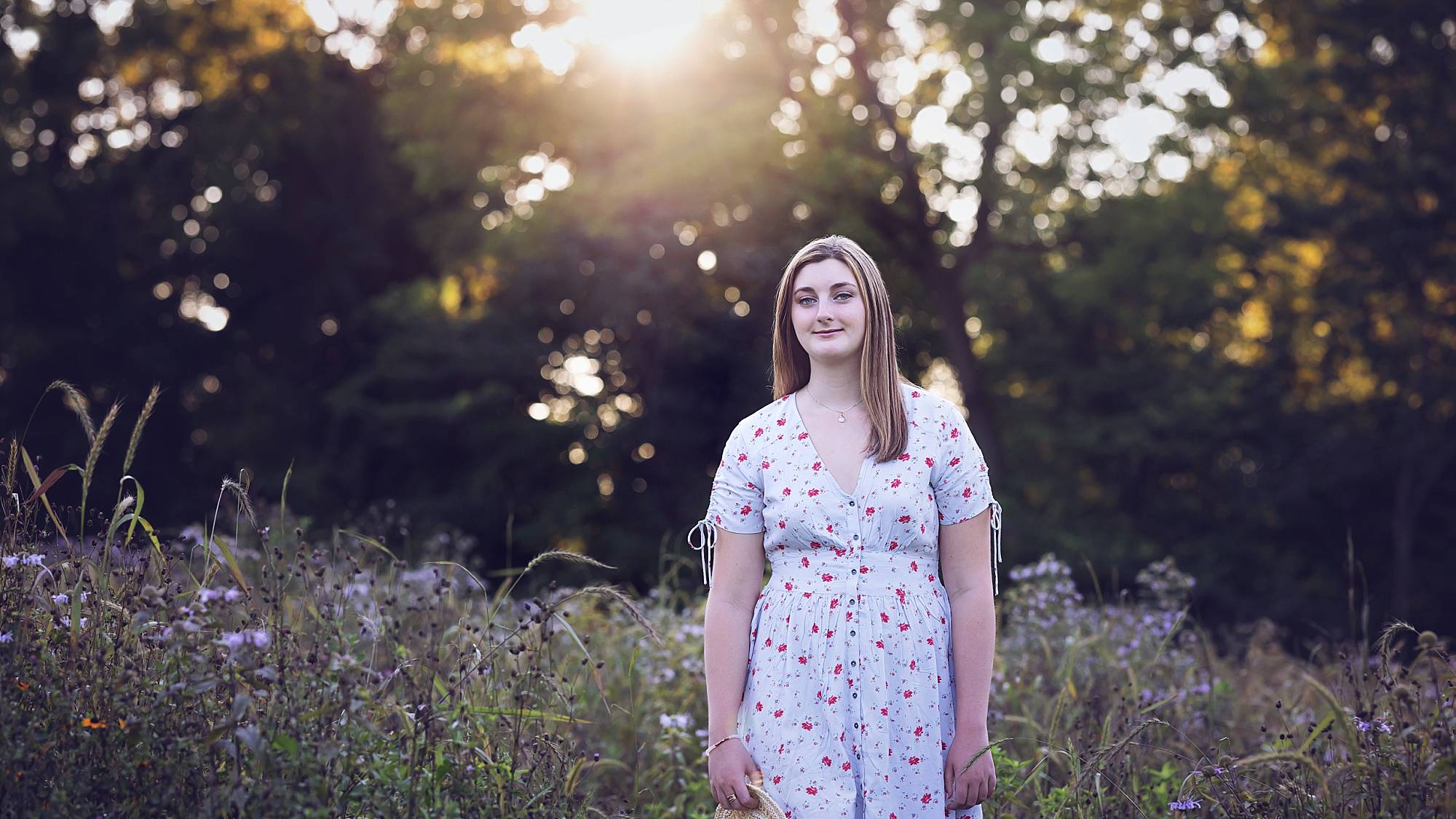 image of a senior girl in a light flower dress. She's standing in a field of wildflowers with the sun setting behind her