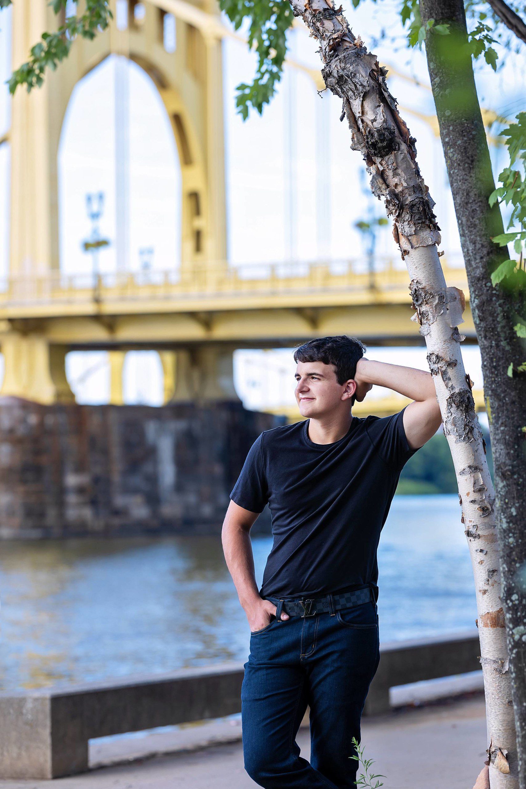 image of a senior guy wearing a black t-shirt and jeans. He has his hand up resting on his head and is leaning against a tree. The yellow Roberto Clemente bridge is in the background