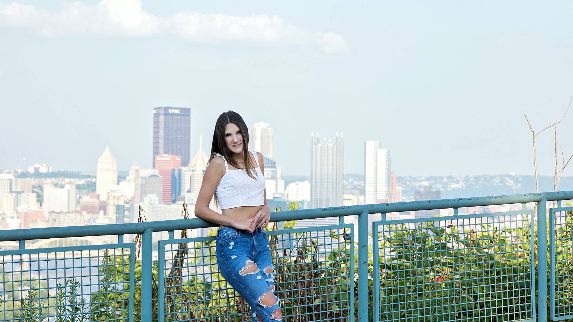 an image from the West End Overlook of asenior girl wearing a white crop top and ripped jeans leaning against a blue railing. The Pittsburgh skyline is behind her.