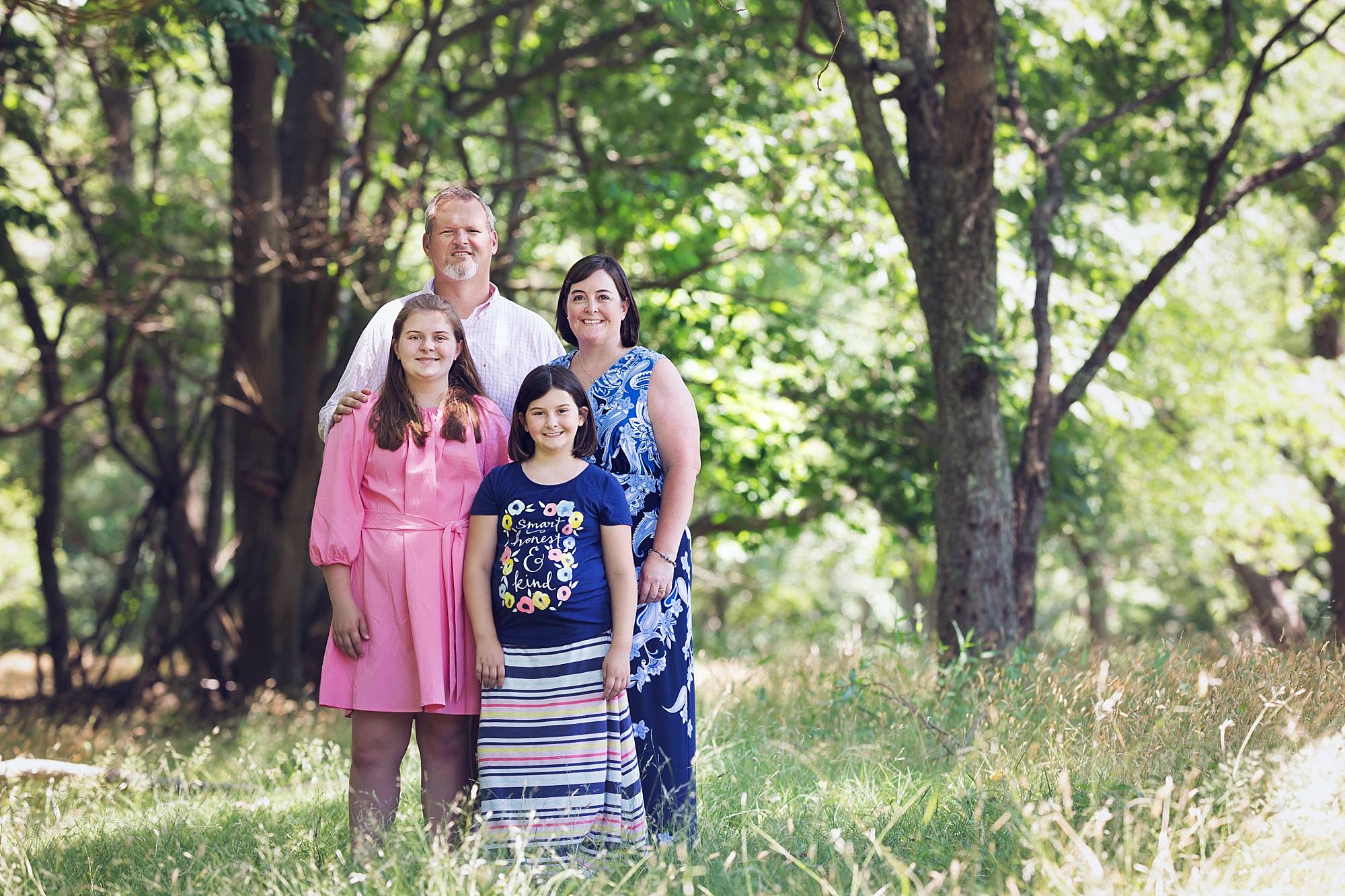 image of a family standing in a clearing with tall grasses and trees. Mom and dad with two daughters, one teenaged and wearing a pink dress. the other daughter is about 10 and wears a blue t-shirt and long skirt.