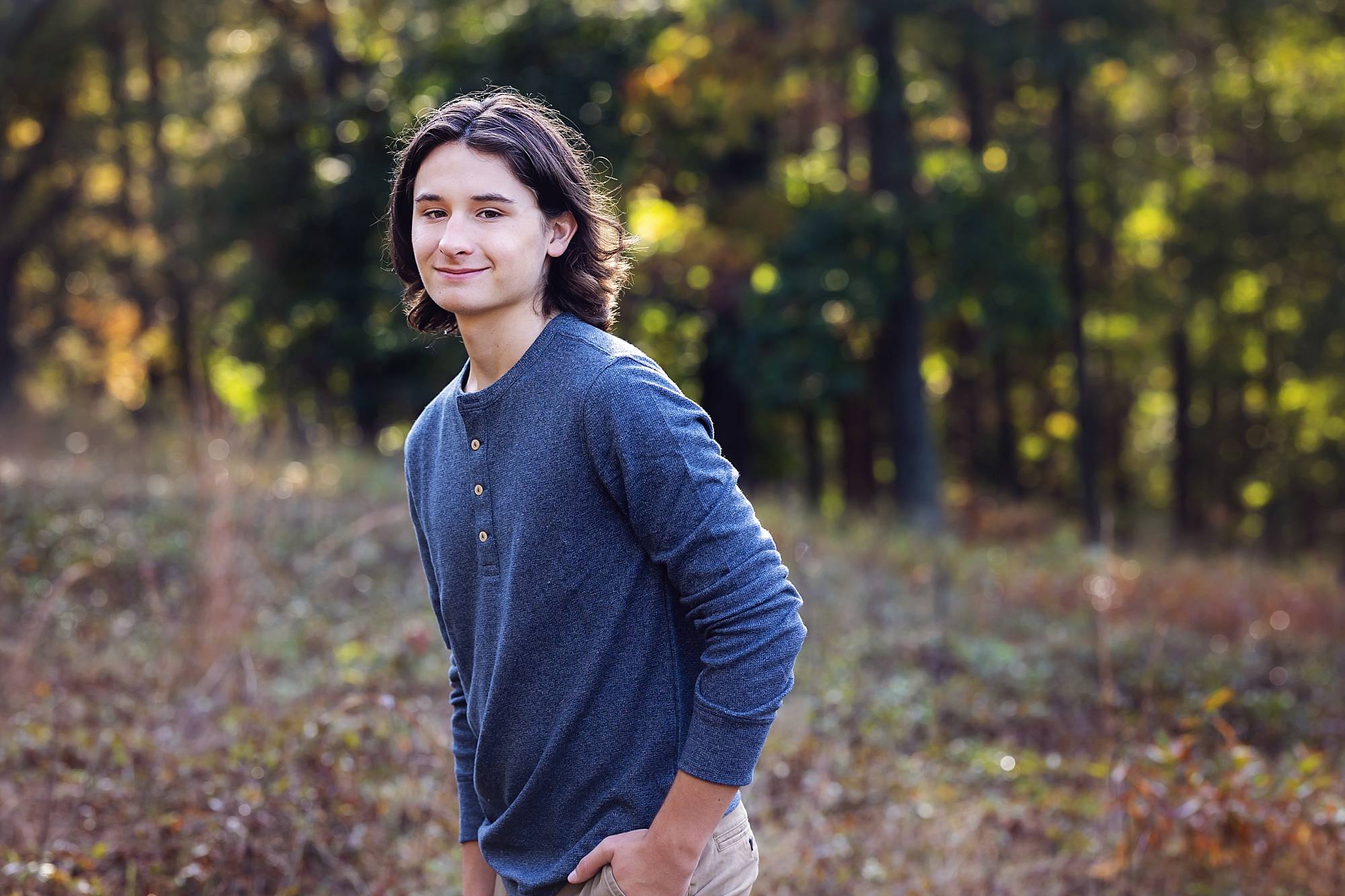 image of a senior guy in a blue henley shirt. he has shoulder length dark wavy hair and is standing in a clearing with a line of trees behind him.
