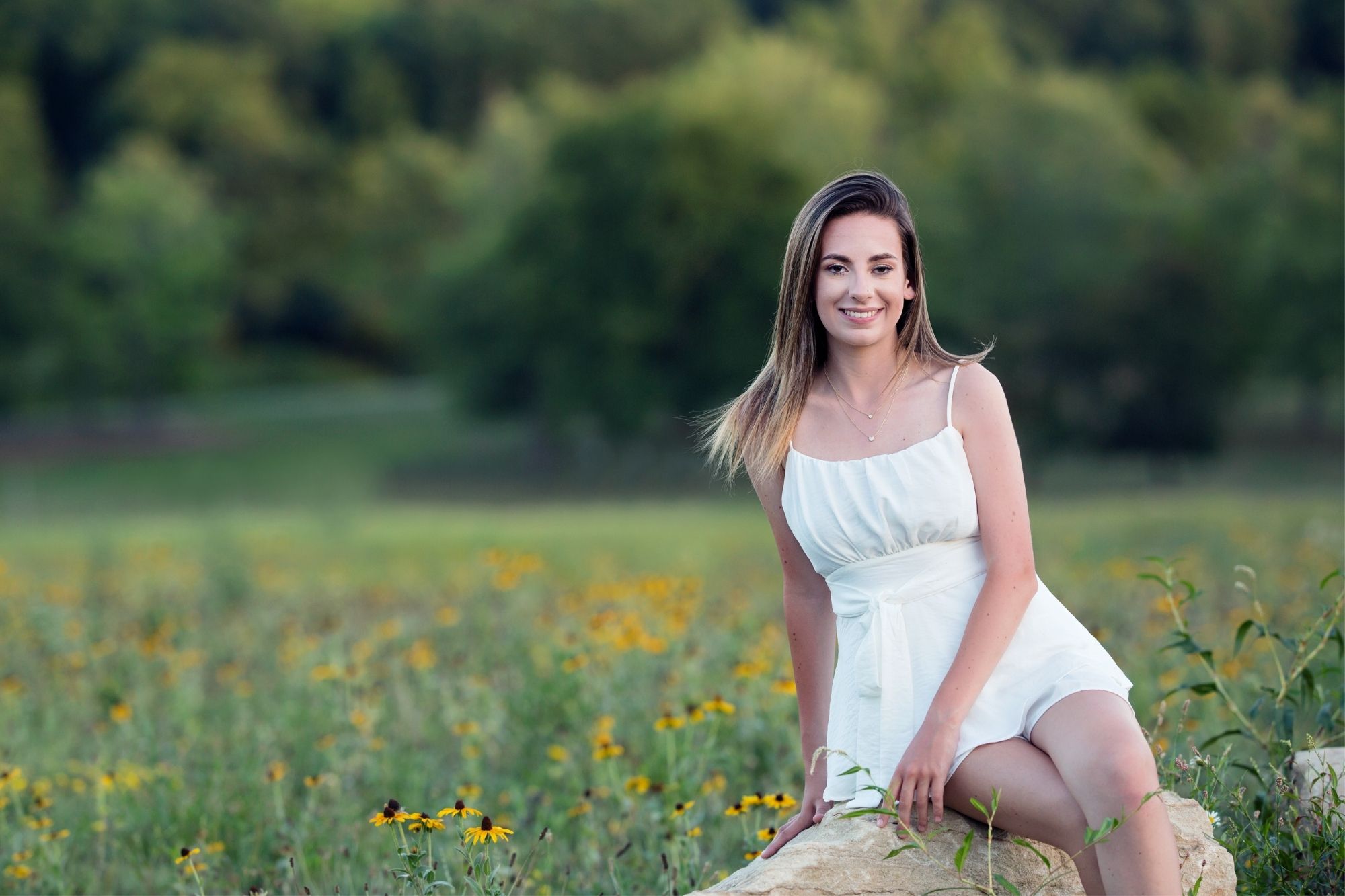 image of a senior girl in a white sundress. she's sitting on a large boulder with a field of black-eyed susans around her