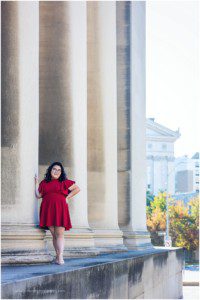 image of a senior girl wearing a red dress. She has dark, wavy hair and glasses and she's standing in front of the Mellon columns