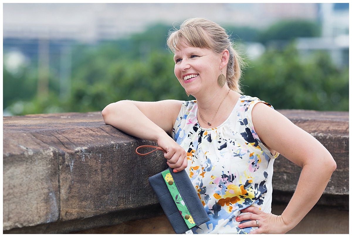 image of Theresa Grosh, owner of Naptime Inspirations, handmade upcycled sewn gifts based in the Lancaster area of Pennsylvania. She's leaning against a stone wall in Schenley Park and holding one of her created clutches.