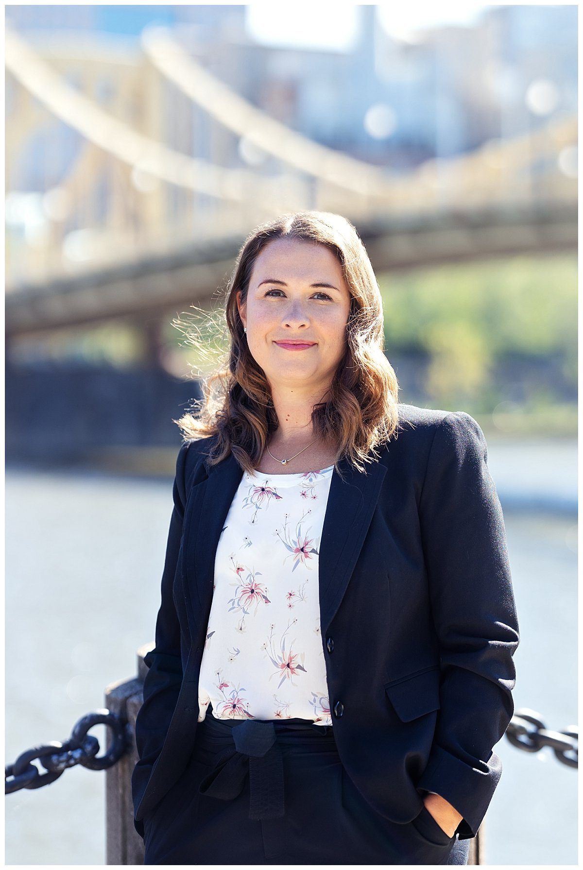 image of a professional woman. She's standing on the North Shore trail in Pittsburgh. The Clemente Bridge is in the background. She's wearing a white blouse with a black blazer.