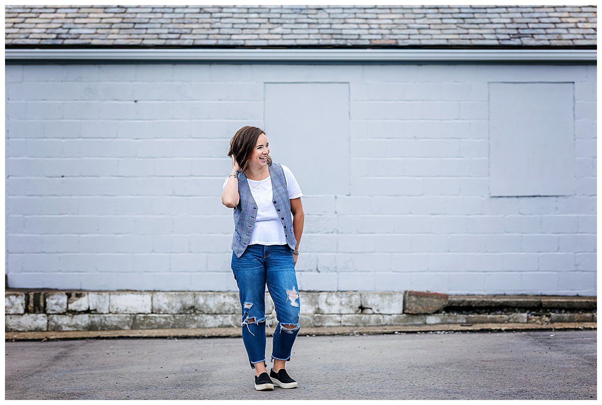 image of a woman with short dark hair. She's wearing a white t-shirt and vest with blue jeans. She's standing in front of a garage in an empty parking lot.