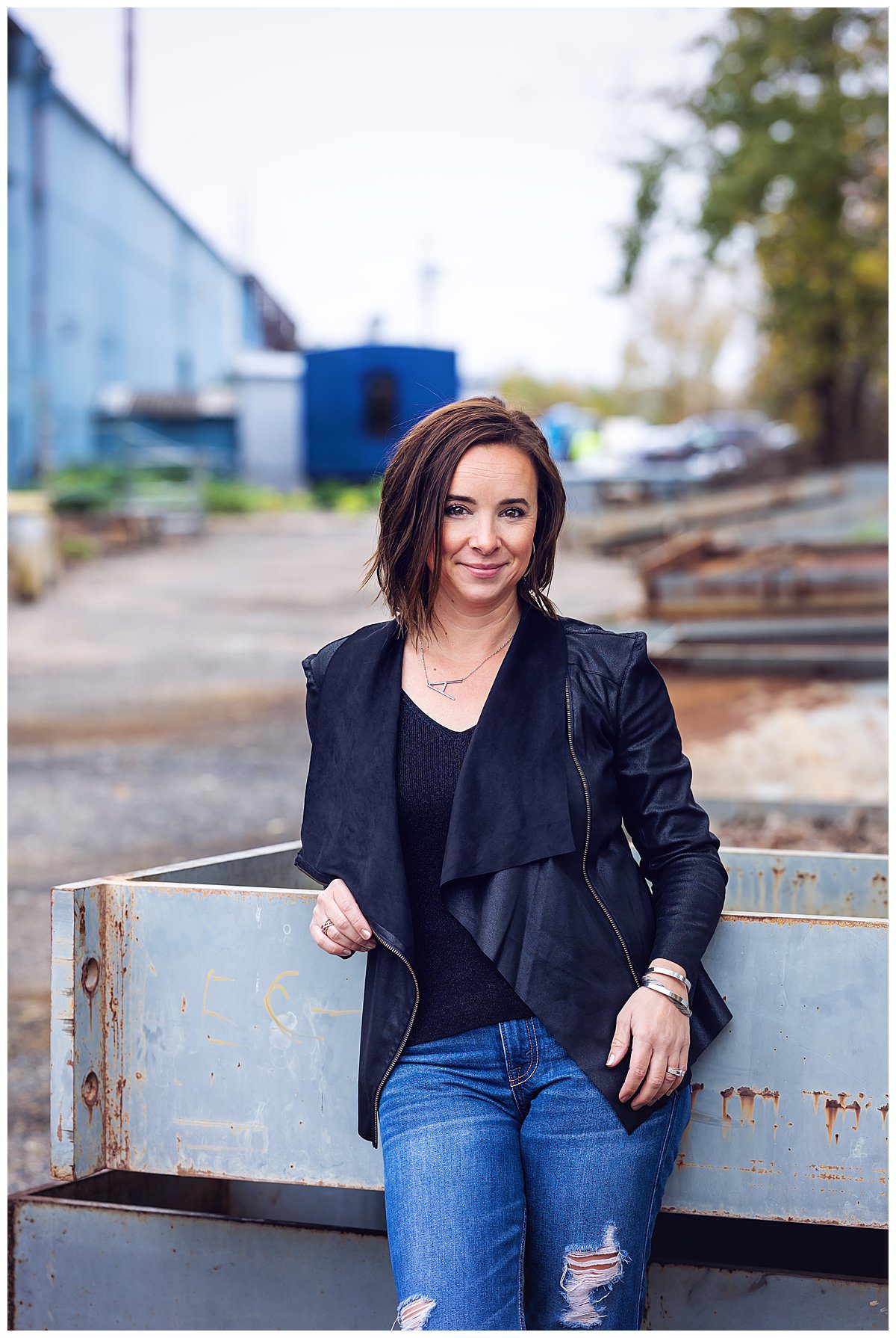 image of Amanda Brisco, photographer. She has short dark hair and is wearing a black jacket with jeans and standing in an industrial alley in Ambridge.