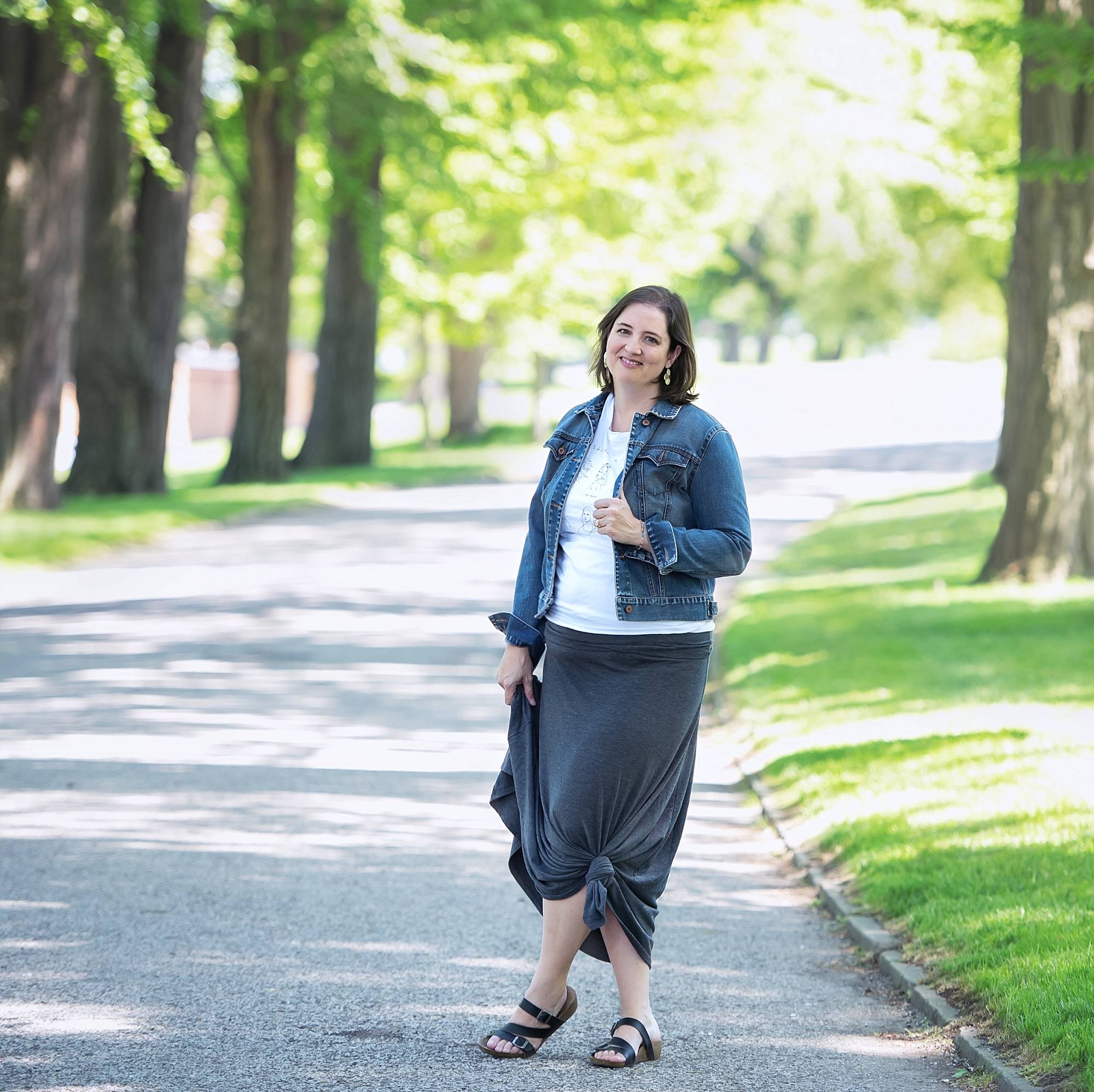 image of Jodi Walsh of Jodi Walsh Photography. She has short dark hair. She's wearing a denim jacket with a white shirt and long gray skirt. She's standing in a road through a park in Pittsburgh.