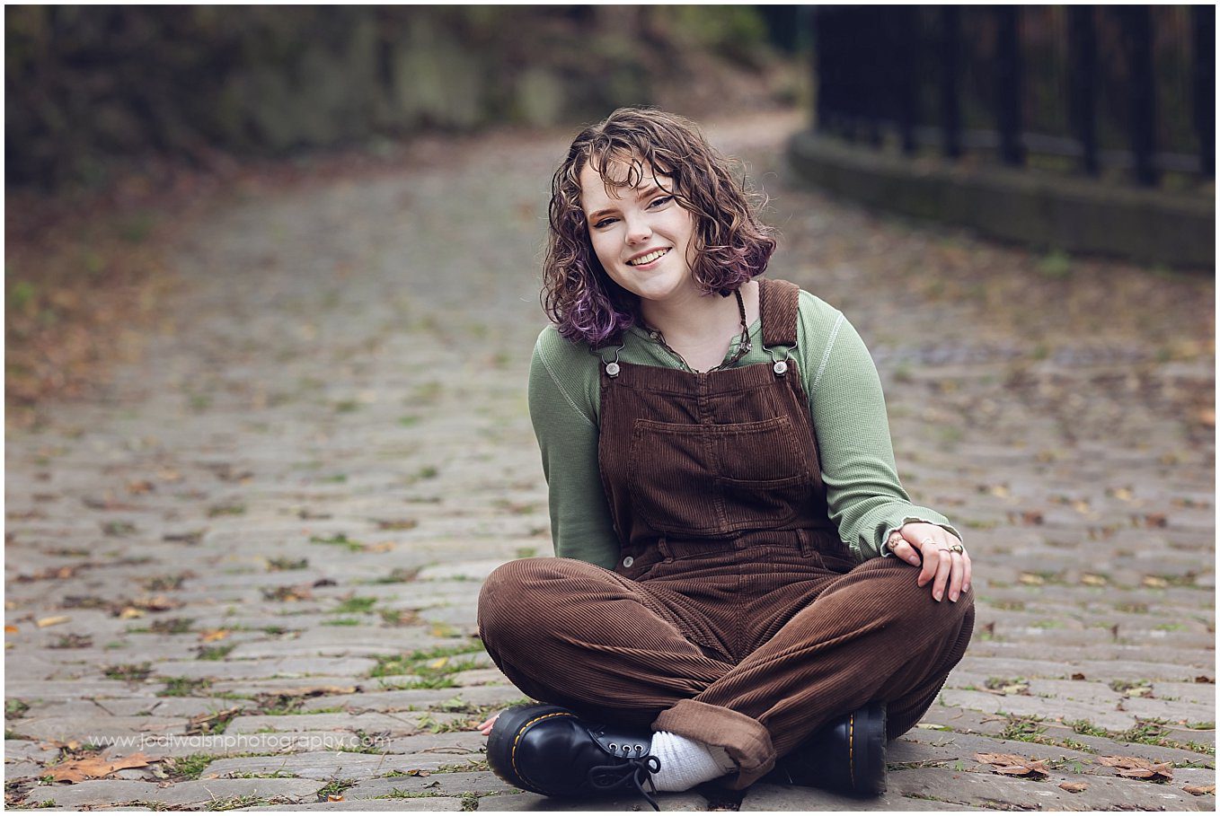 senior girl wearing brown overalls and an olive green top. She's sitting with crossed legs on a cobblestone street in Schenley Park, Pittsburgh