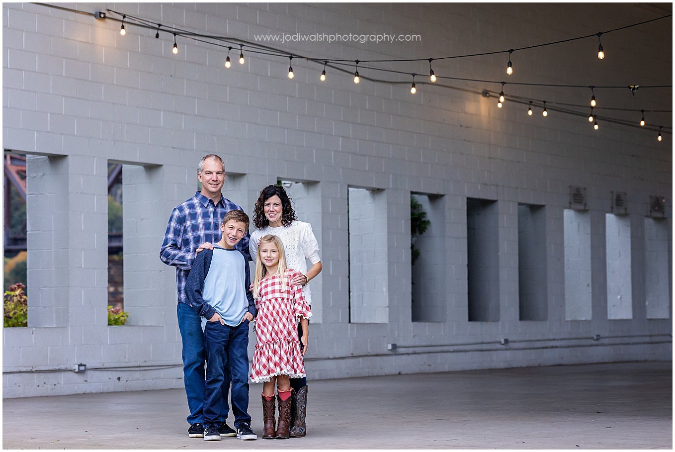 image of a family (mom, dad, boy and girl) standing in a stone pavilion with twinkle lights strung across the ceiling in Allegheny RiverTrail Park.