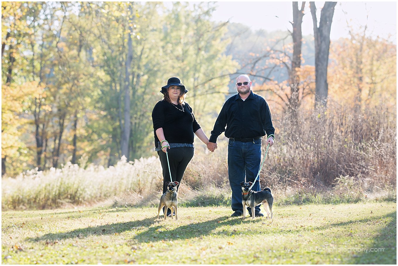 image of a couple wearing black. They're holding hands and standing in front of a field of tall grasses with two little dogs on leashes in front of them.