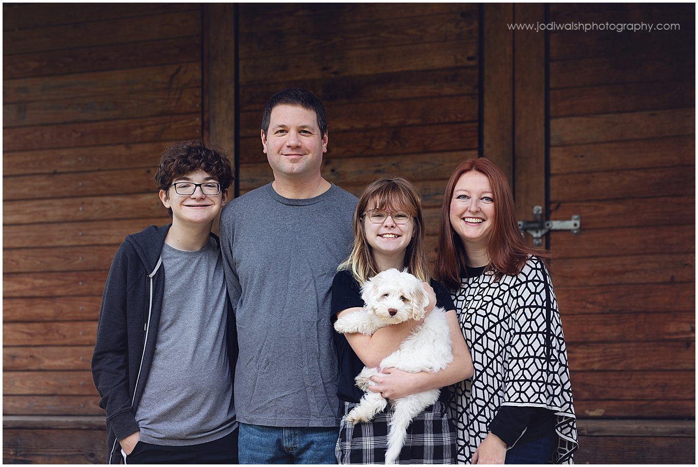 image of a family (mom, dad, teen son and daughter) with their puppy standing in front of a wooden building at Boyce Mayview Park.