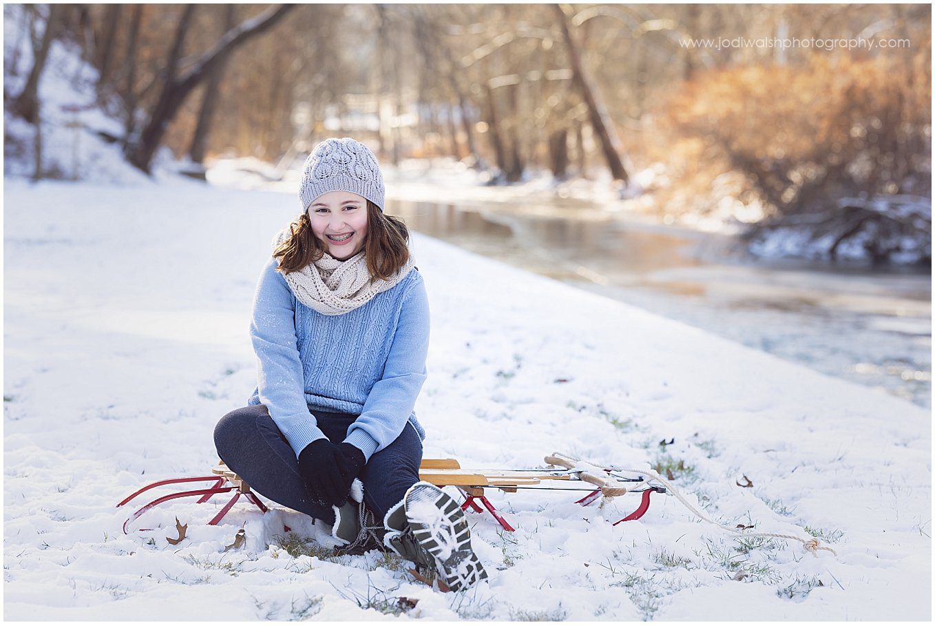 image of a teen girl sitting on a sled in the snow with a frozen creek behind her. She's wearing a light blue hat and sweater.