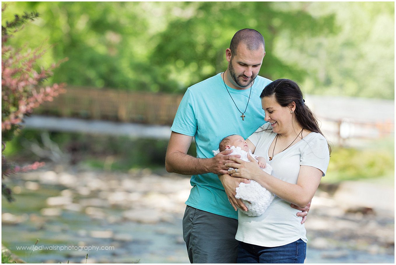 image of a young couple with a newborn. They're standing together and looking down at the baby in mom's arms. There is a creek and a footbridge in the background.