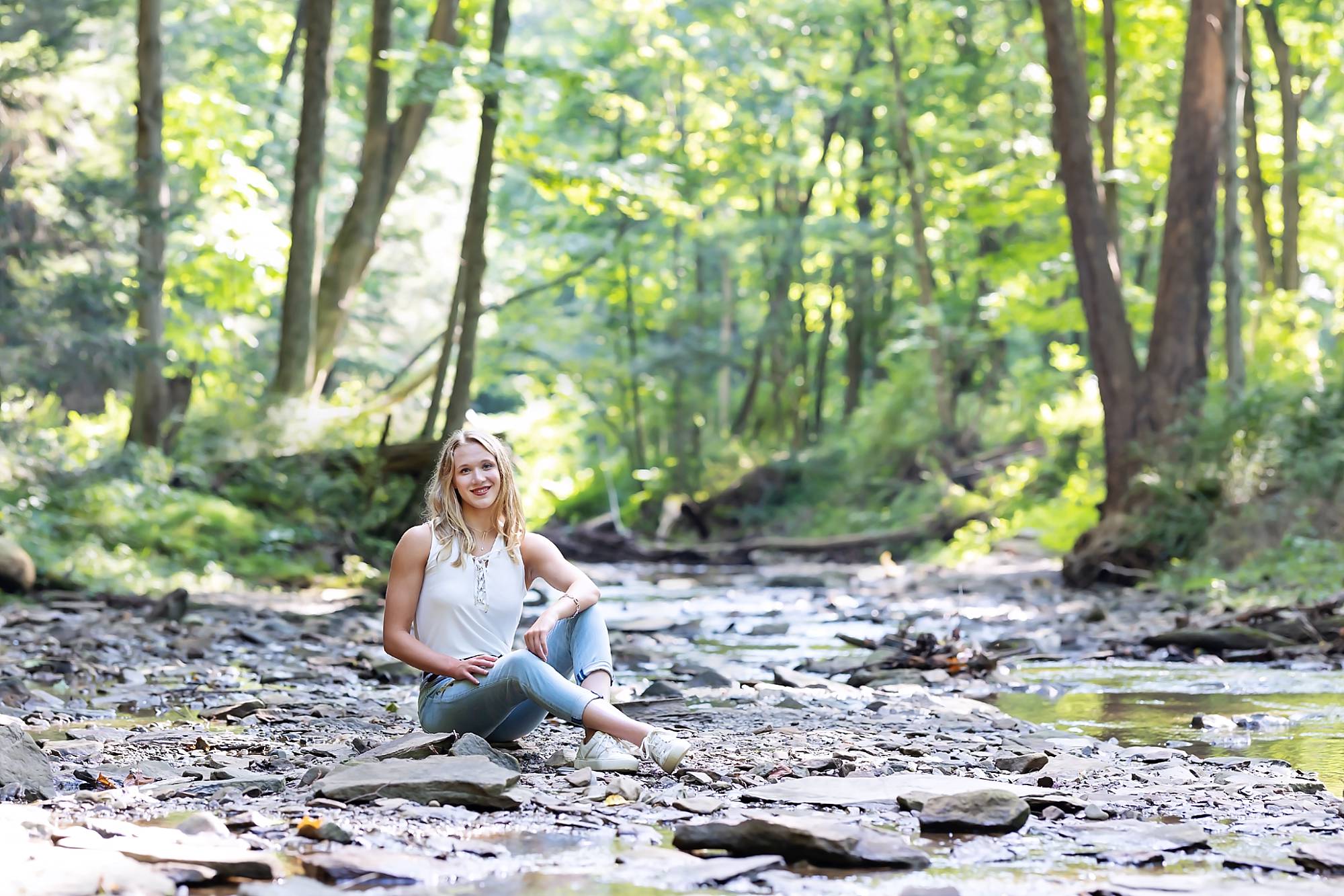 image of a senior girl wearing a white top and jeans sitting on a rock in a shallow creek bed.