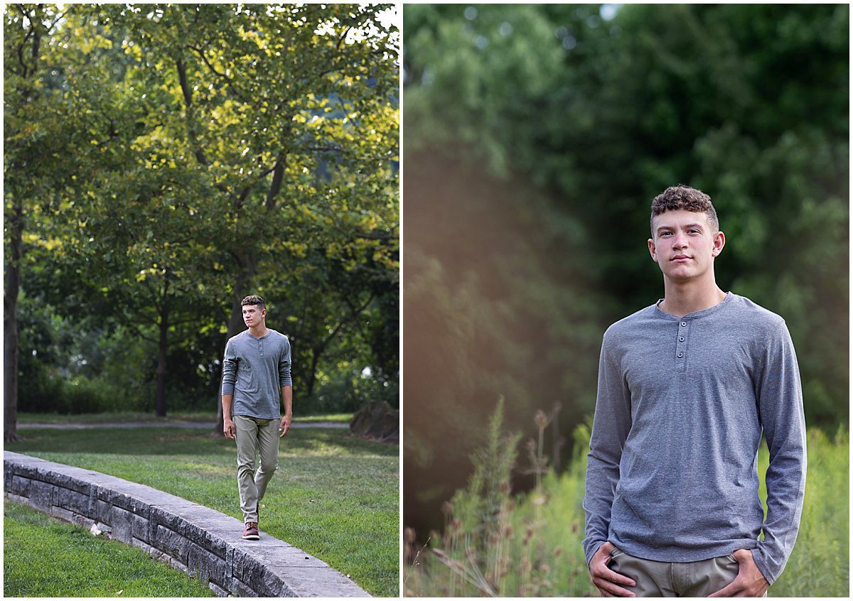 images of a senior guy at a park on Herrs Island. In one image he's walking along a short stone wall and looking off to the left. In the second image he's standing in tall grasses with pine trees behind him. He's wearing a gray henley shirt and khaki pants.