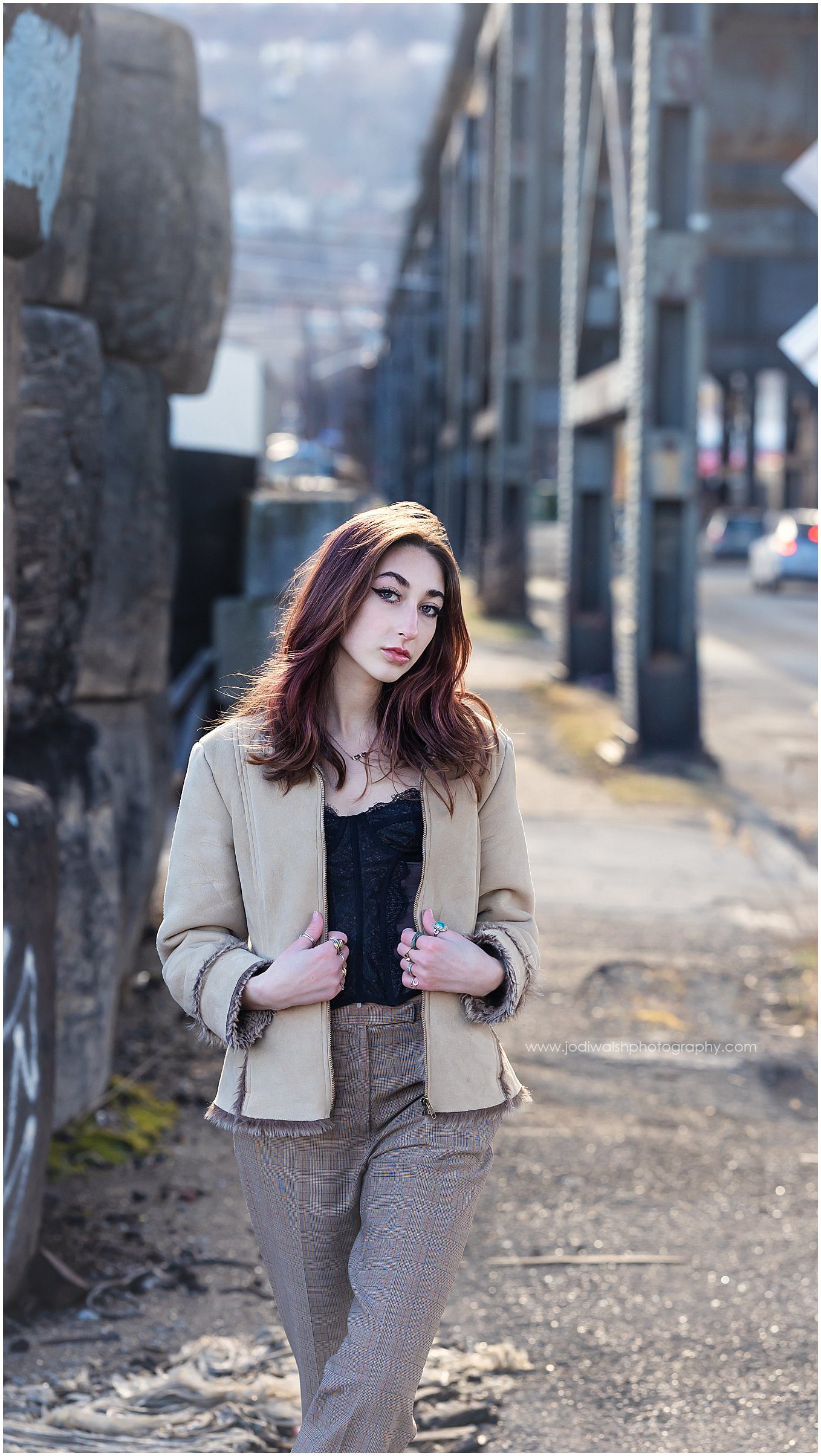 image of a senior girl walking along an industrial alley in Pittsburgh's Strip District. She has long dark hair and is wearing beige plaid pants and a beige jacket with a fur collar.