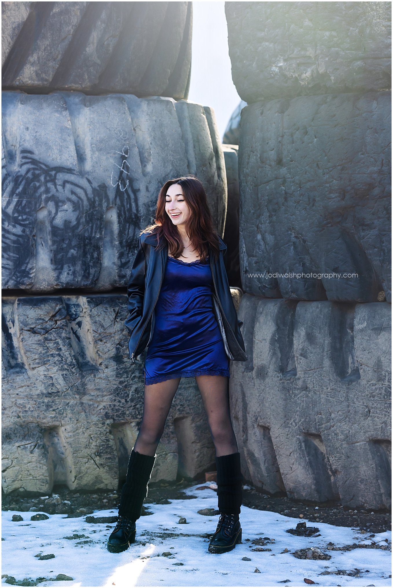 image of a senior girl wearing a short blue silk dress, black leather jacket and black boots. She's standing in front of stacked industrial tires along an alley in Pittsburgh's Strip District. She's smiling and looking away with her eyes closed.