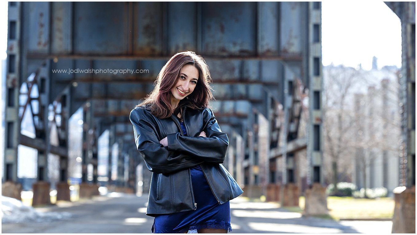 image of a senior girl standing in the street with a large train trestle overhead. She's wearing a black leather jacket and has her arms crossed but with a big smile.