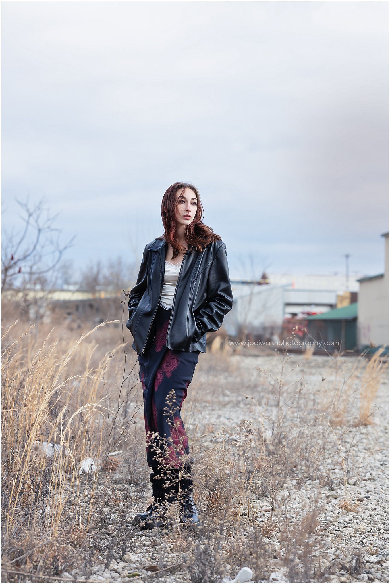 image of a senior girl standing in an empty alley with a stretch of tall grasses off to the left side. There's a row of warehouses in the distance. She's wearing a long deep red and black skirt and a black leather jacket.