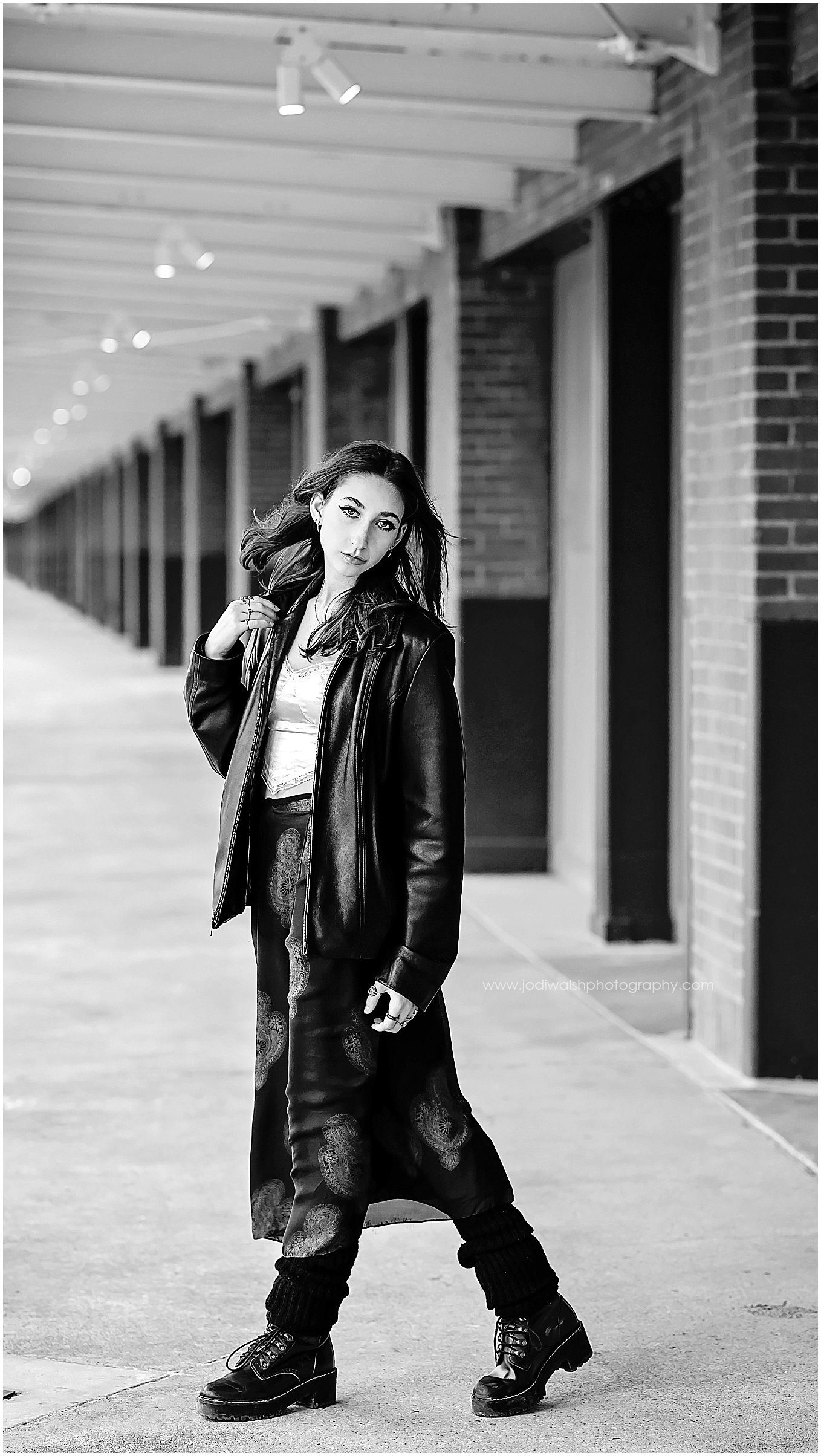 black and white image of a senior girl standing along the back walkway of the Terminal Building in Pittsburgh's Strip District. She's wearing a black leather jacket, a long dark skirt, and combat boots.
