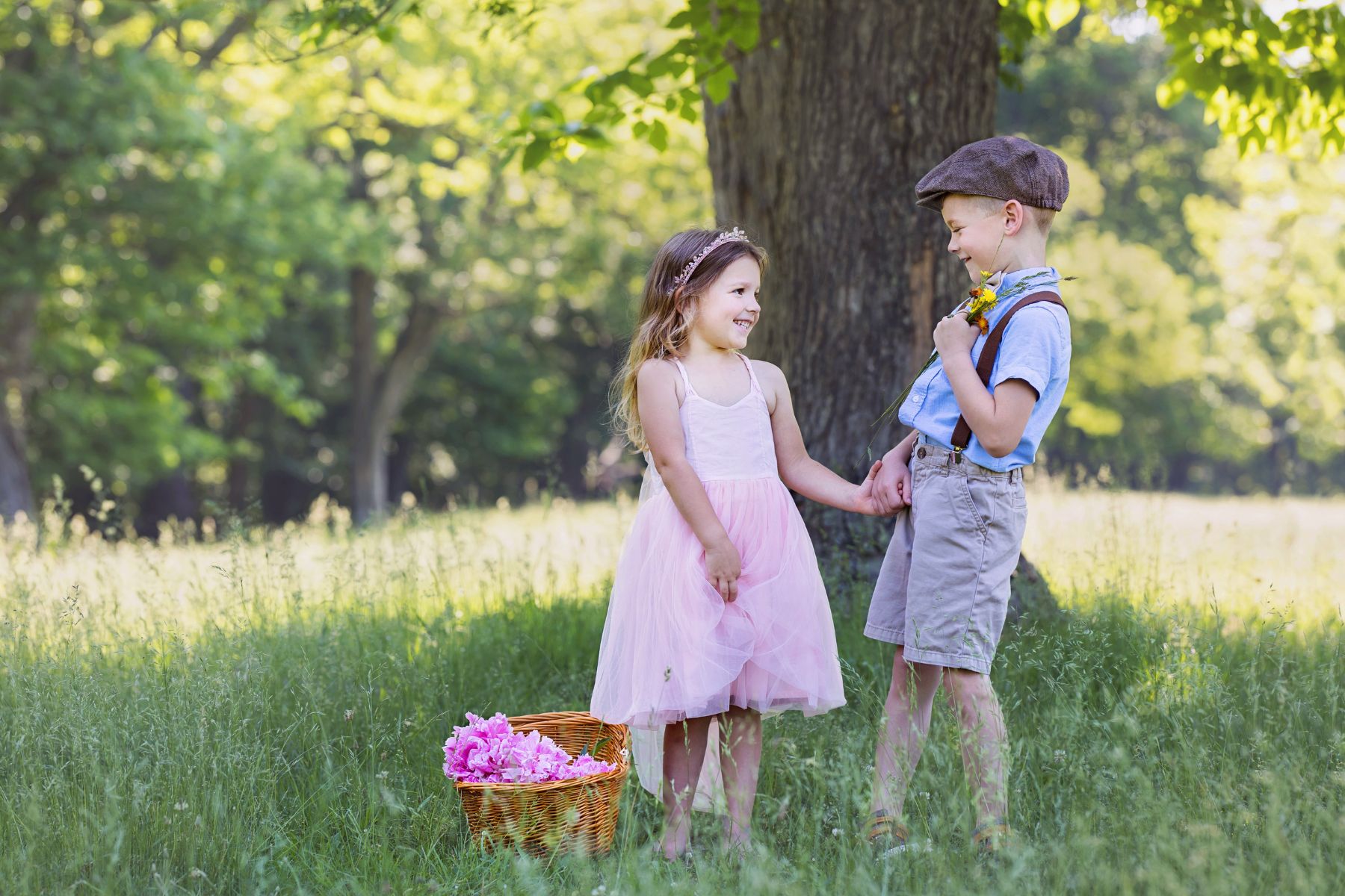 image of a littel boy and girl, brother and sister. They're standing in the tall grass in the shade of a big tree. She has a basket of pink flowers at her feet. He's wearing a newsboy cap and suspenders. They're holding hands as they look at each other.