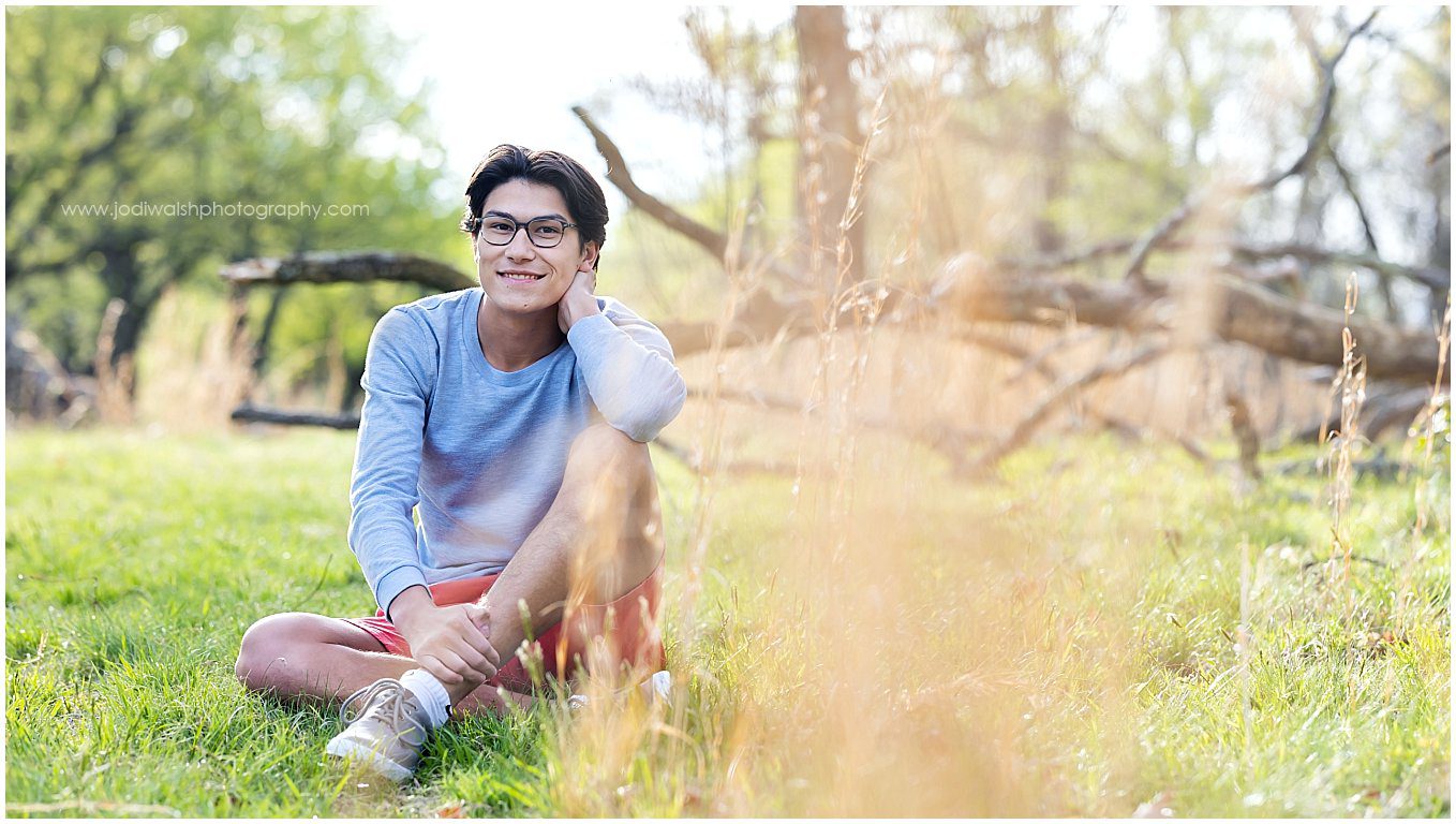 image of a senior guy with dark hair and glasses. He's wearing a light sweater and shorts and sitting in a field with tall grasses. There is a fallen tree in the distance behind him.