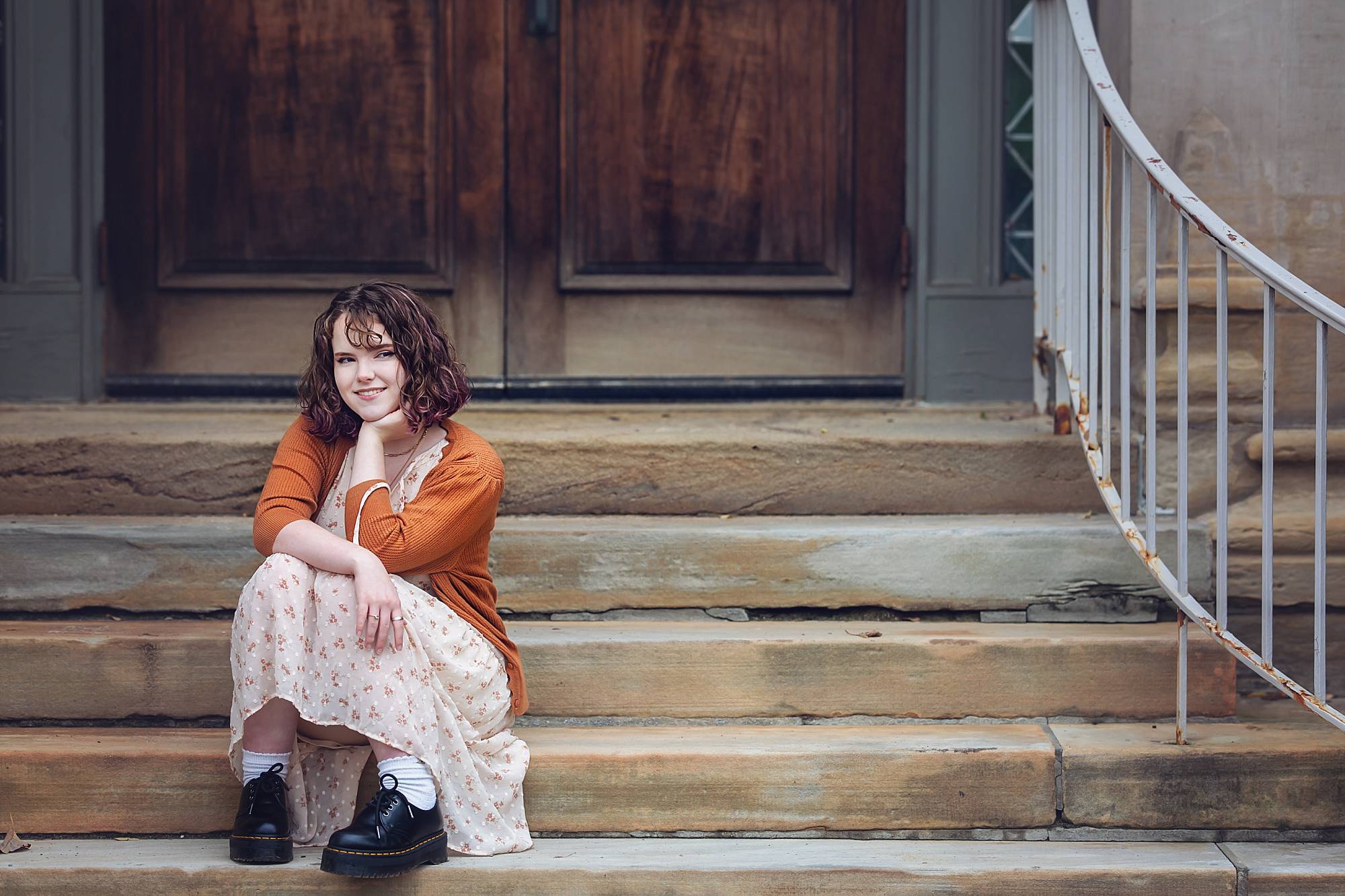 image of a senior girl sitting on stone steps near Phipps Conservatory in Pittsburgh. She's wearing a flowered long dress, with an orange knit sweater and black combat boots.