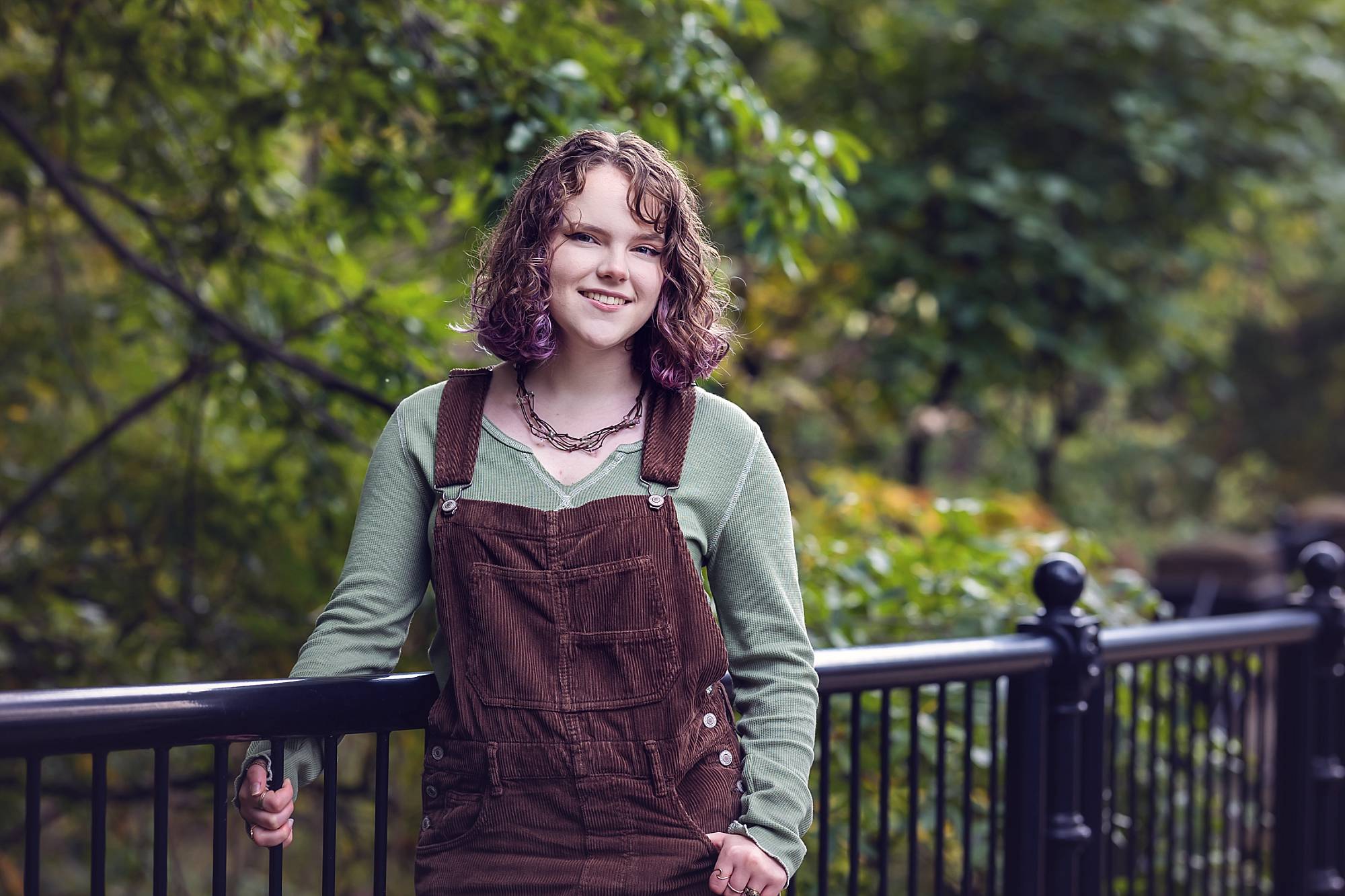 image of a senior girl standing by a black iron fence in Schenley Park, Pittsburgh. She has brown, shoulder length, curly hair and is wearing brown overalls with a green top.