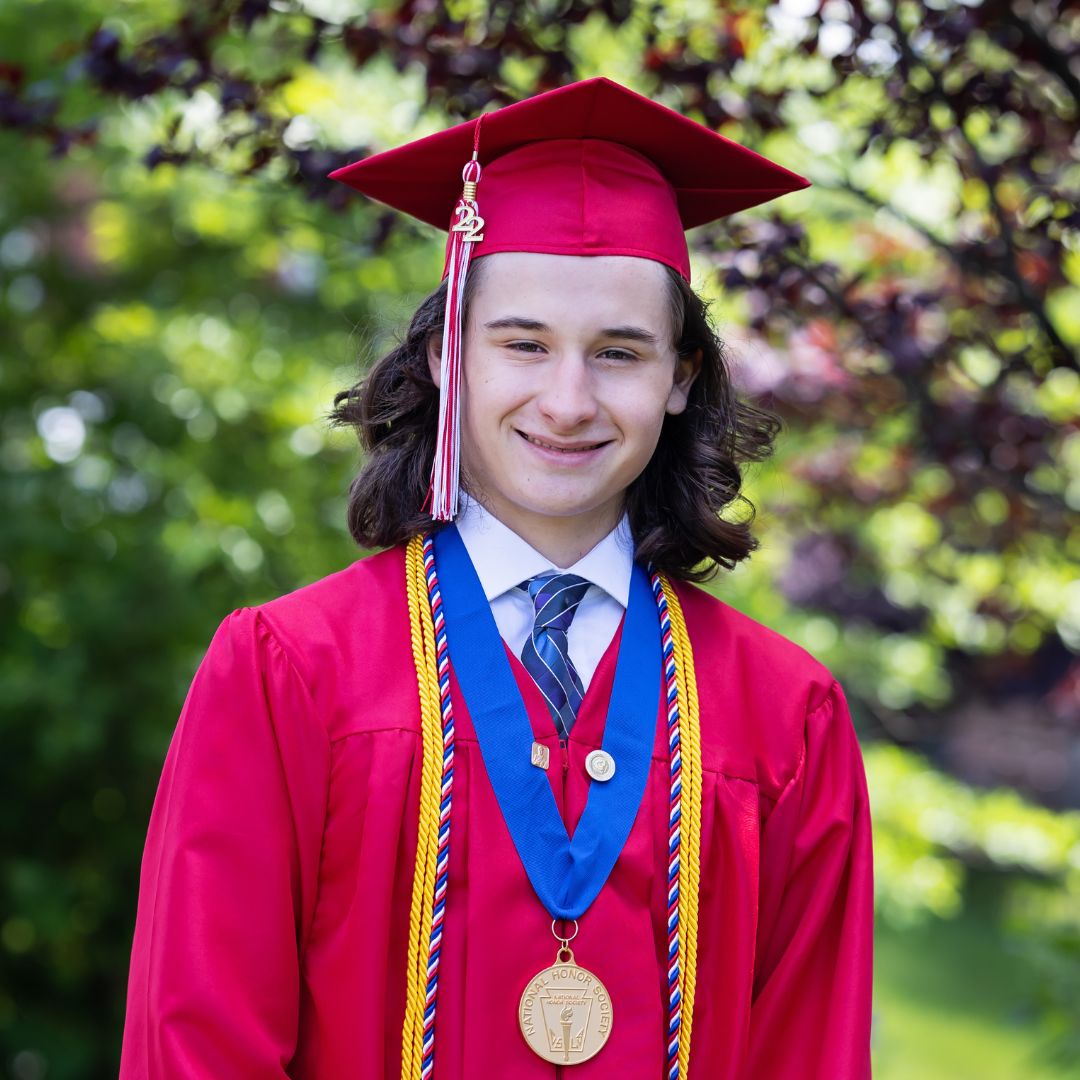 image of a high school senior guy. He's wearing a red cap and gown with gold medal on a blue ribbon and a gold cord. He has dark hair that is shoulder length and wavy. There are trees in the background.
