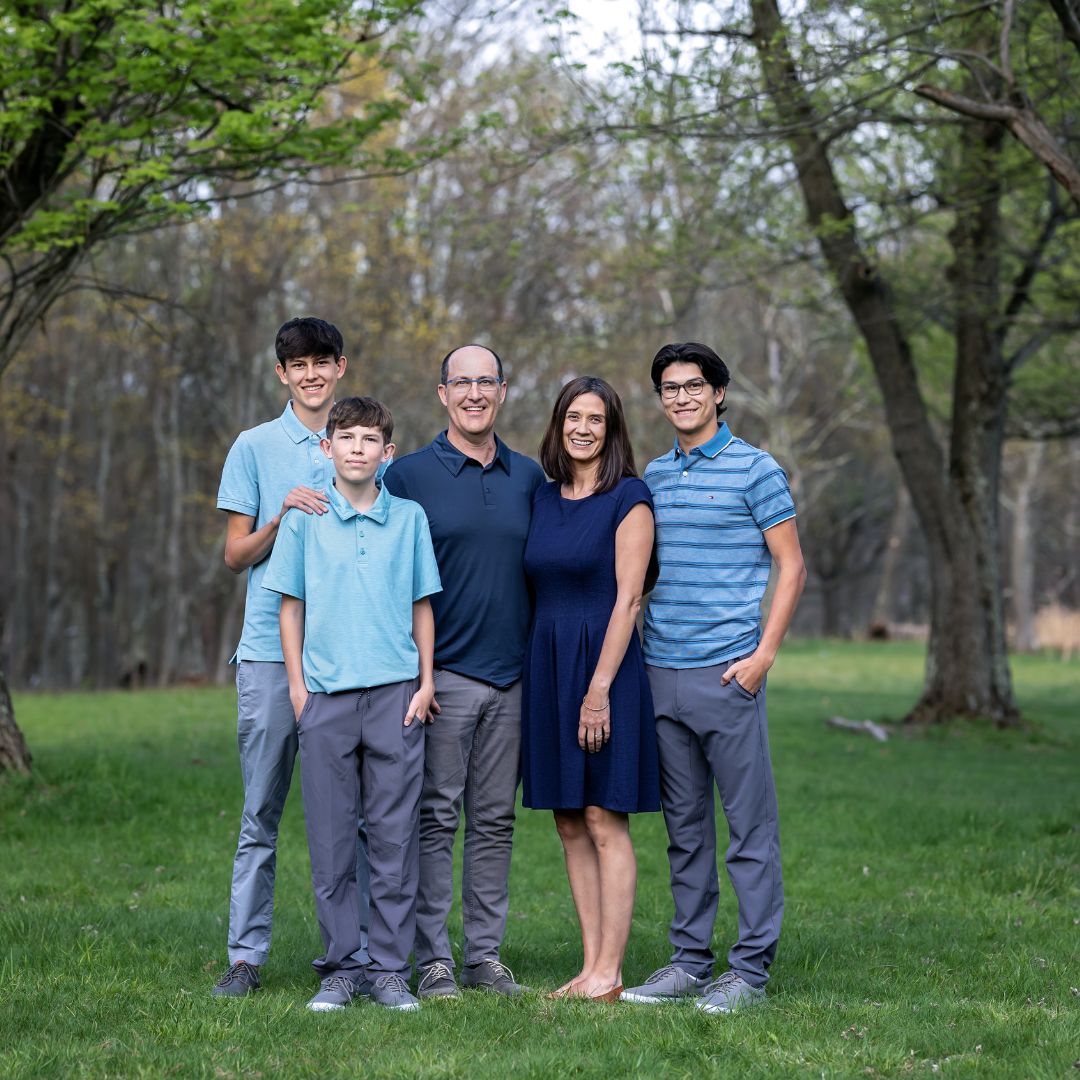 image of a family of five standing in a clearing with woods in the background. Mom and dad stand in the center with 3 older boys around them. Two of the boys are tall and in high school. They are all wearing shades of blue.