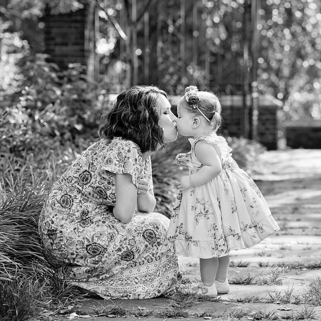 black and white image of a mother kissing her toddler girl. They're both wearing flowered dresses and they're in a garden at Mellon Park in Pittsburgh.