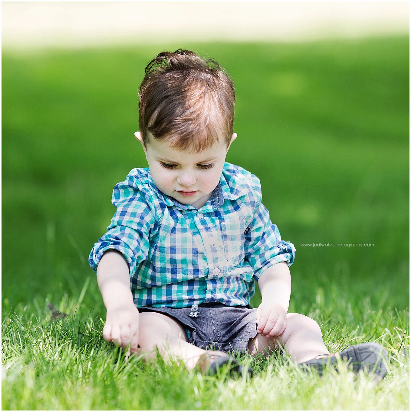 image of a little boy sitting in the grass. He's looking down at the blades of grass. He's wearing a blue plaid shirt and navy shorts.